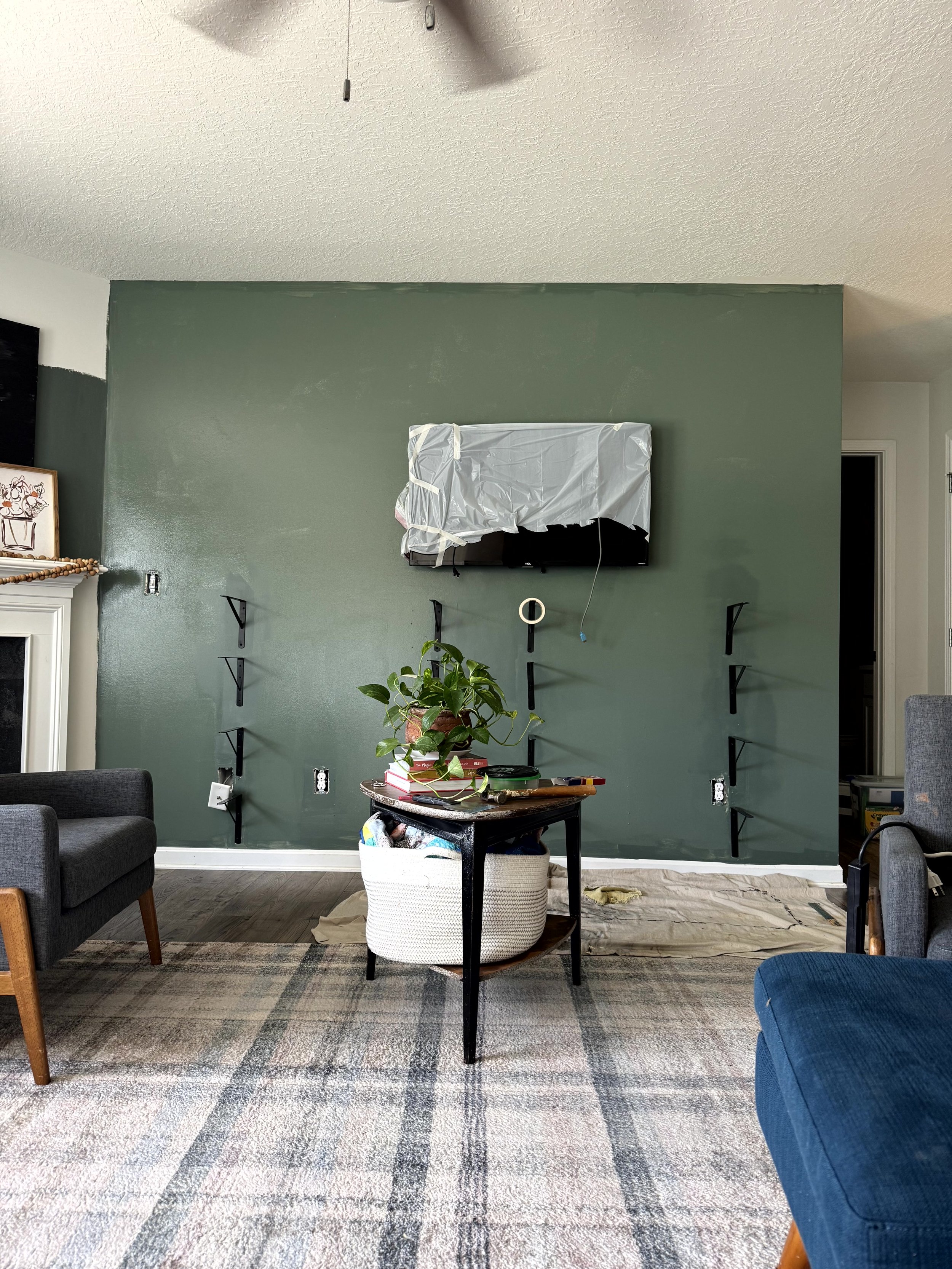  A living room wall fully painted green, but no shelves on the brackets. The TV is still covered and the baseboard is still white. The decision to drench the trim has not yet been made.  