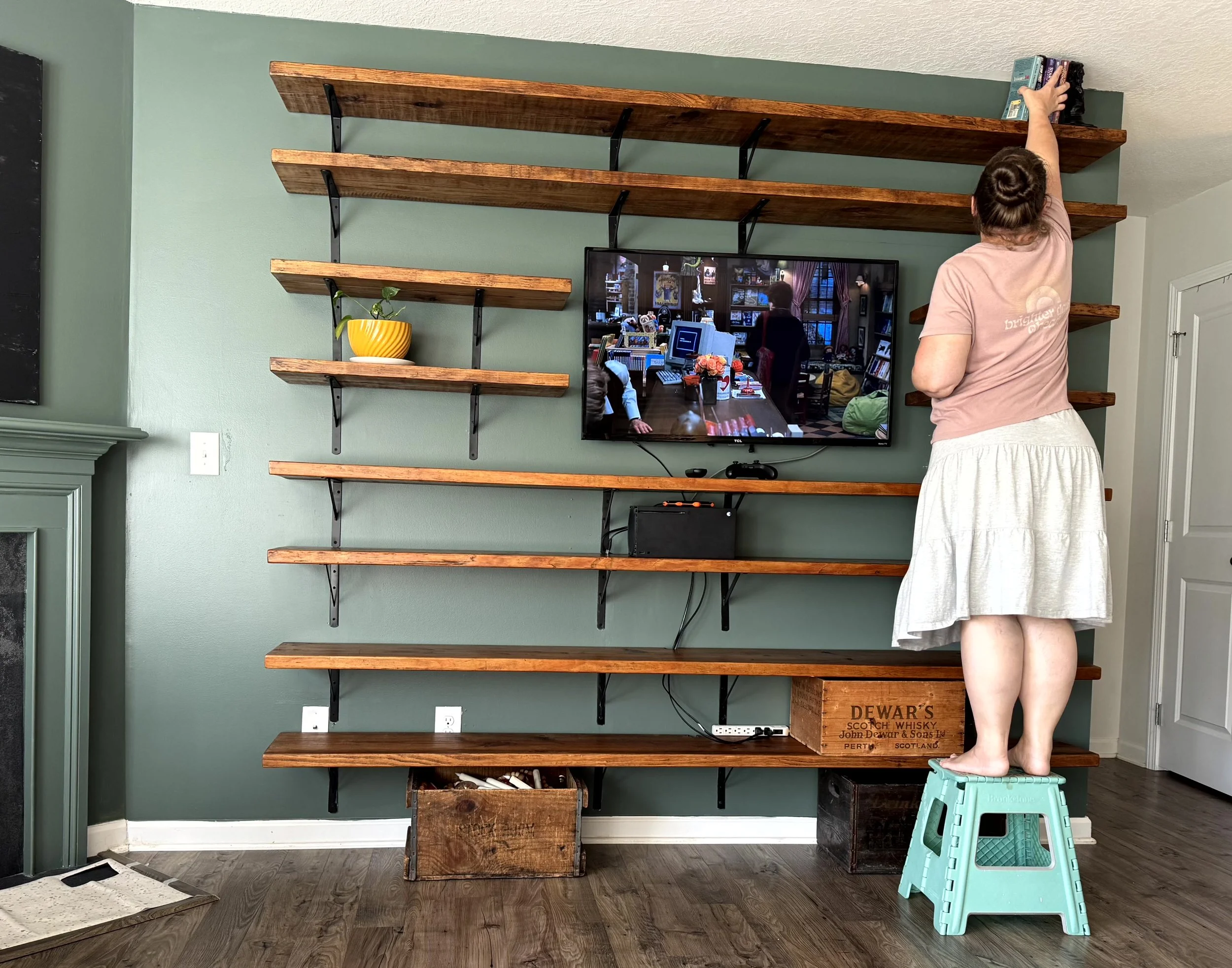  A cozy green living room, warm wood shelves surroundng  a tv mounted on the wall. a woman is standing on a teal step stool putting books up on the top shelf of the shelves that have obviously been newlly installed  