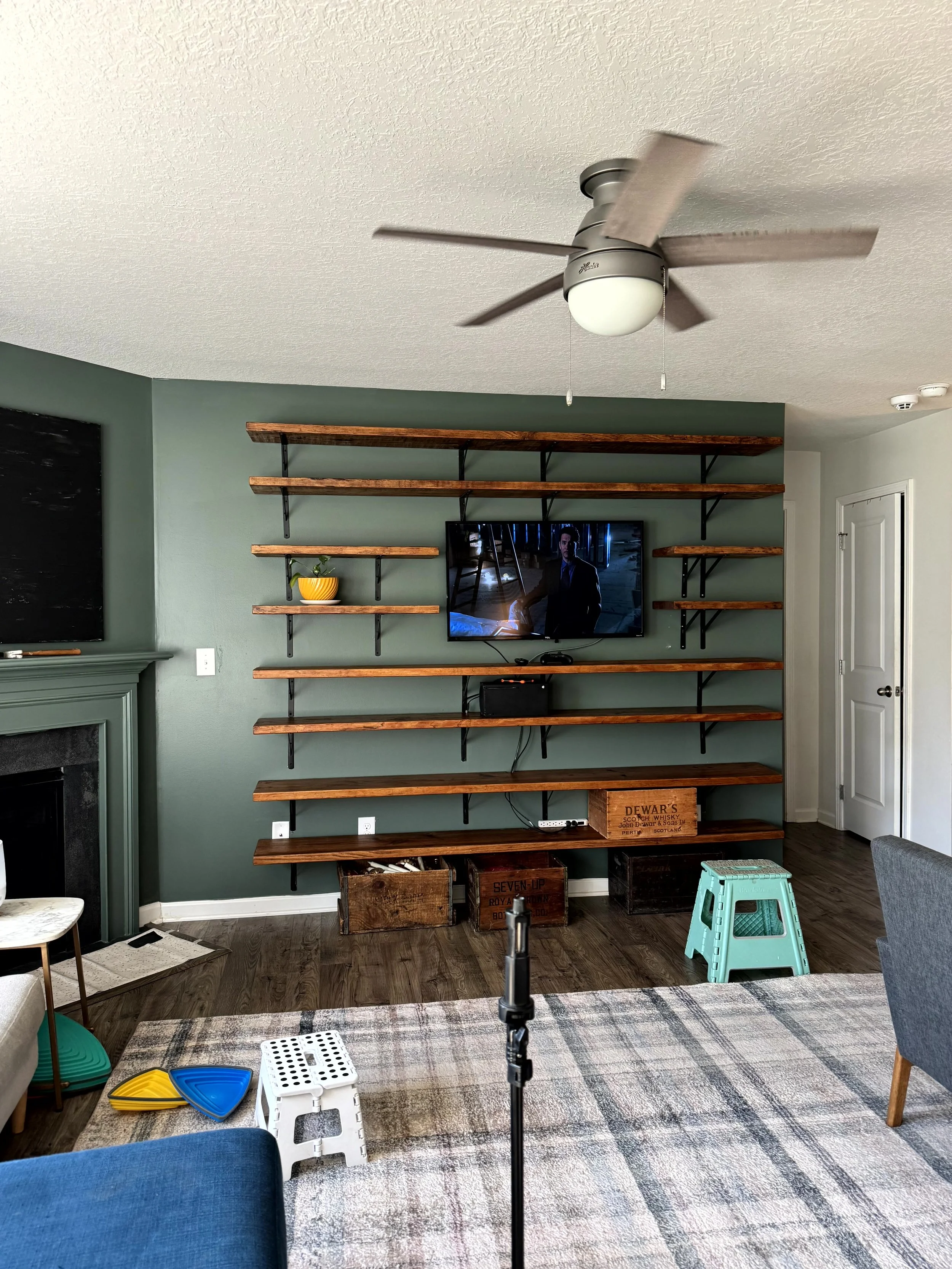  A cozy living room with a warm plaid rug, green paint, a wall of warm wood shelving. The shelves are still empty and the room pulled apart. it is obviously still in process of being put back together after the shelves were installed.  