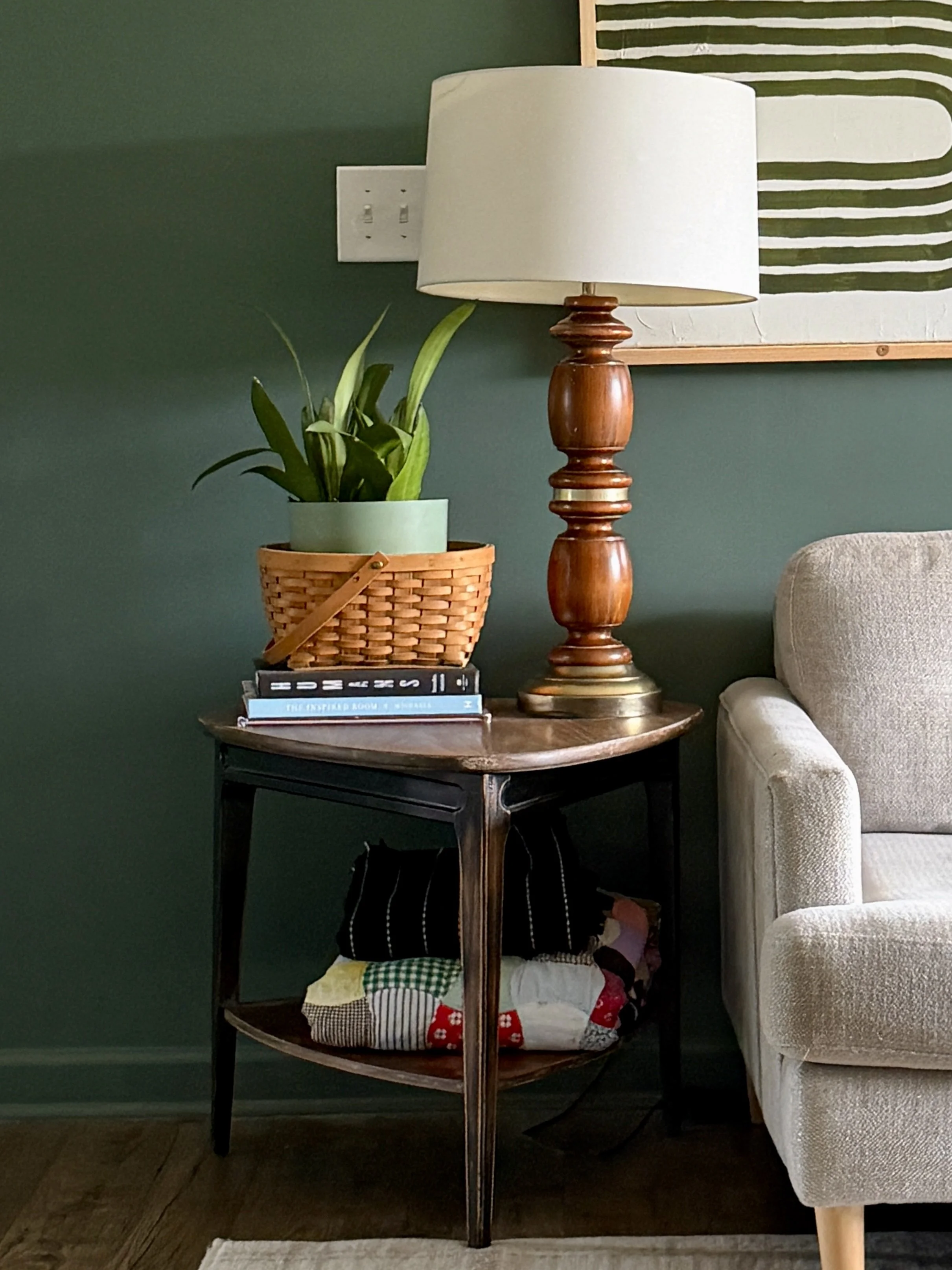  A shot of the table she was working on, it’s far from complete  but it looks great with colorful blankets stacked on the bottom shelf, and a stack of books with a basket with a moonshine snake plant in it and a vintage wood and brass lamp next ot it