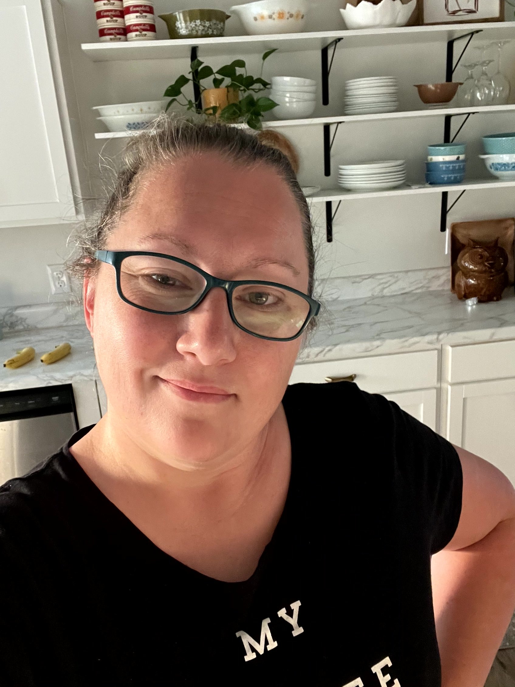  Selfie of woman in black shirt with the temporary counter/cabinets, and white open shelving on the wall with black brackets. 