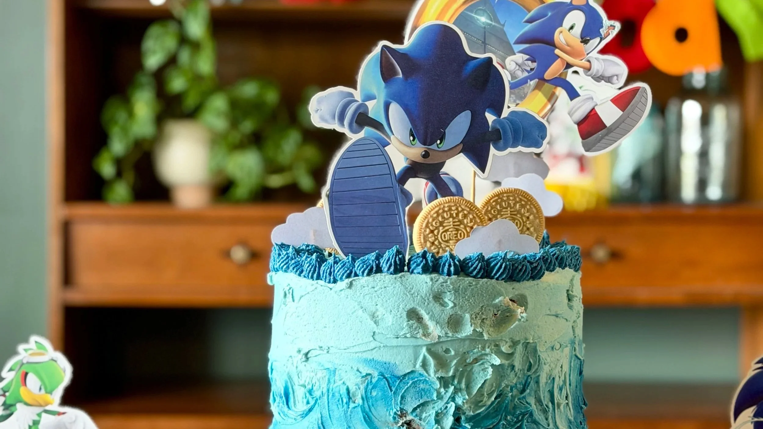 Two toned blue sonic the hedgehog birthday cake. It has gluten free golden oreos as rings and a coupld of large table top decor pieces as the topper. Set against a backdrop of a dining room with vintage wood furniture and gren walls, with plants