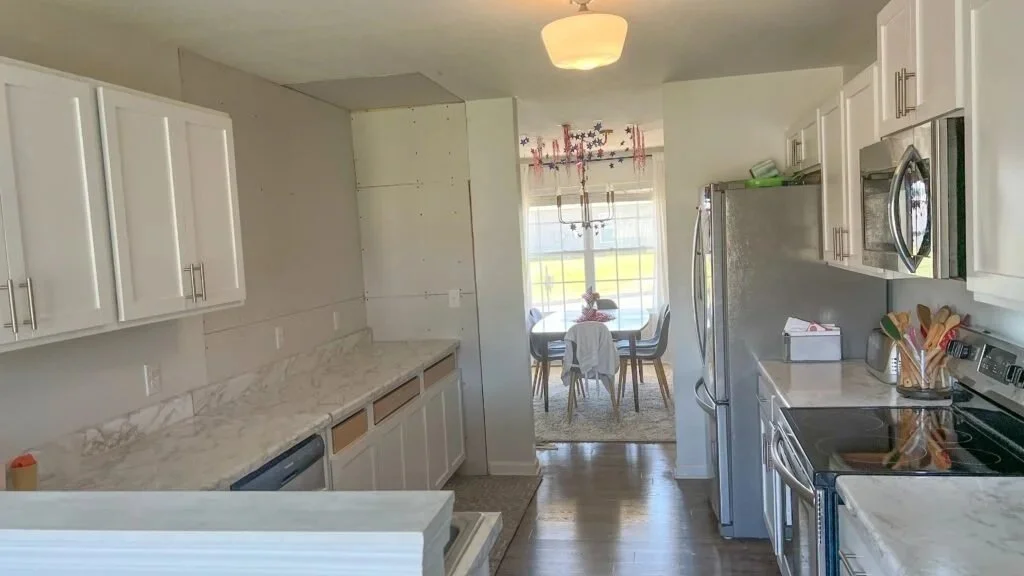 A whte kitchen, mid renovation. part of the walls are just drywall, the cabinets do not all have drawer fronts, the dining room is visible through the doorway