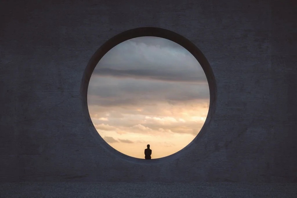 Persona en silueta frente a un círculo en una pared, con cielo nublado y atardecer de fondo.