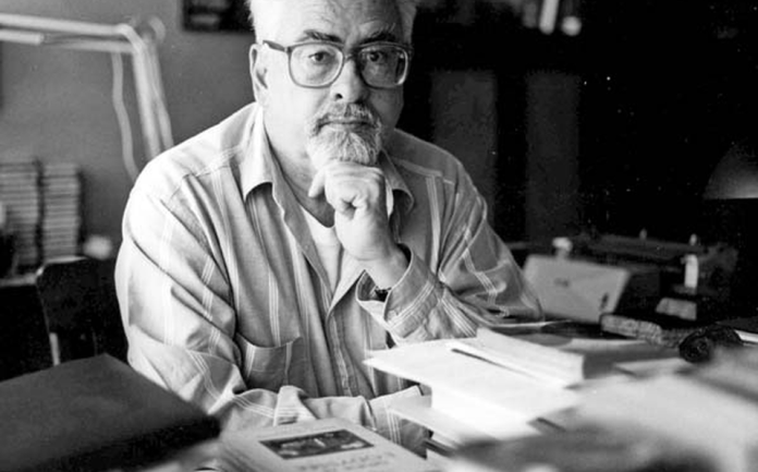 Hombre con gafas en una oficina, rodeado de papeles y libros, en una fotografía en blanco y negro.
