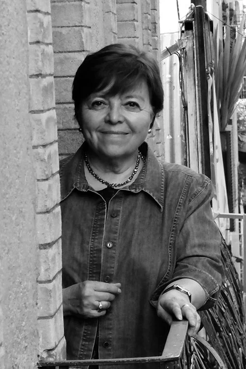 Mujer mayor con cabello corto y vestida con chaqueta de mezclilla, sonriendo y apoyada en un balcón de una casa de ladrillos en un ambiente exterior.