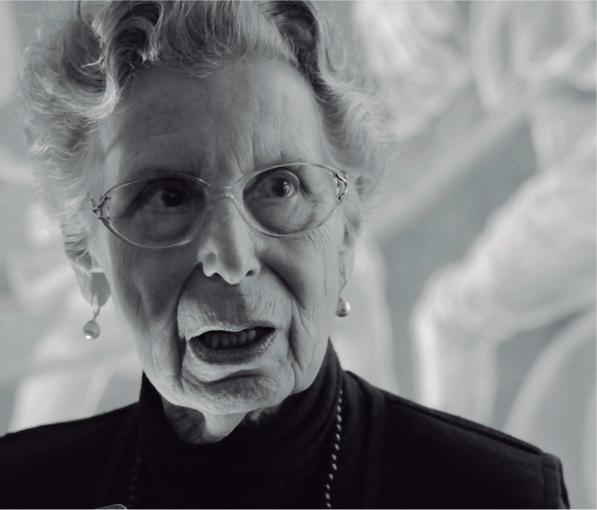 Retrato en blanco y negro de una mujer anciana con gafas, cabello rizado y expresión facial sorprendida o pensativa.
