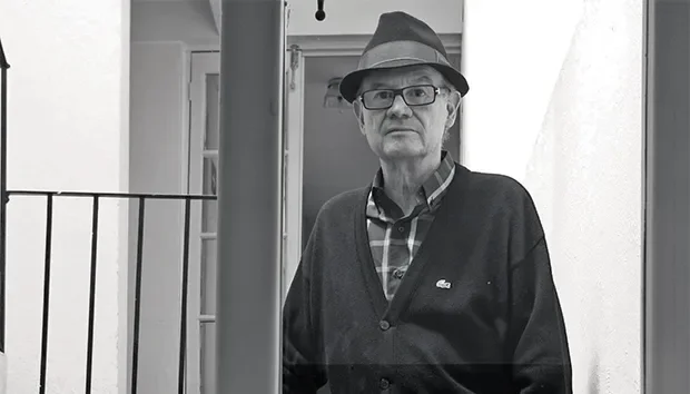 Hombre mayor con gafas, sombrero y suéter oscuro, de pie frente a una puerta en interiores, en una imagen en escala de grises.