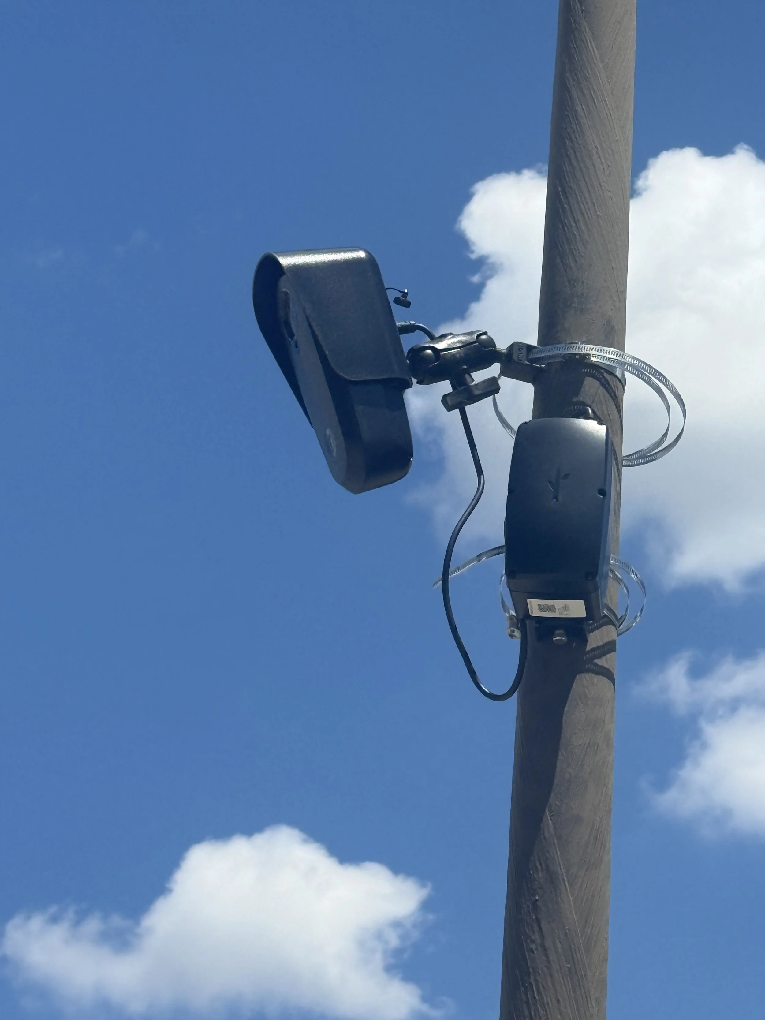 Close-up of a utility pole with a Flock Safety camera and solar panel attached, against a blue sky with scattered clouds.
