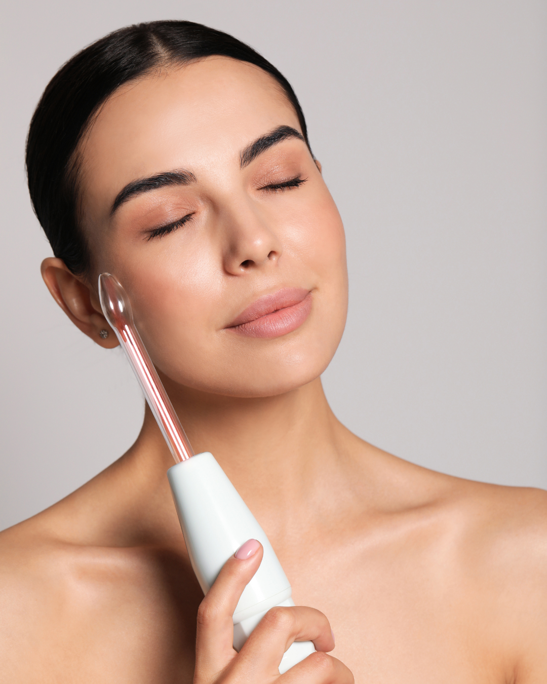 A woman with dark hair and closed eyes uses a facial steamer on her face.