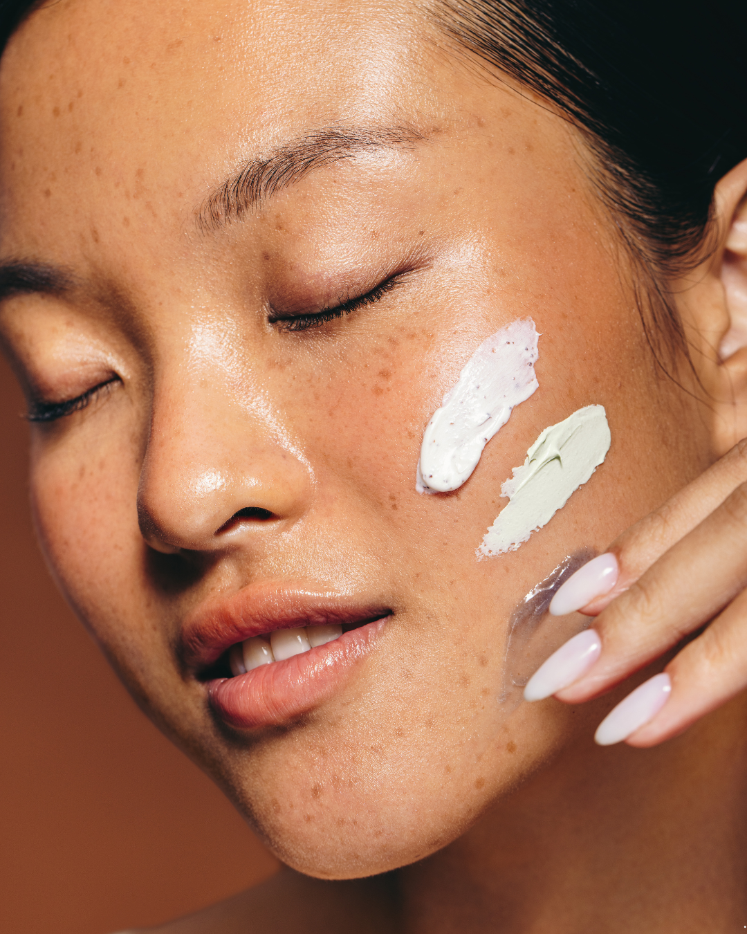 Close-up of a woman's face with closed eyes, showing skincare product swatches on her cheek.