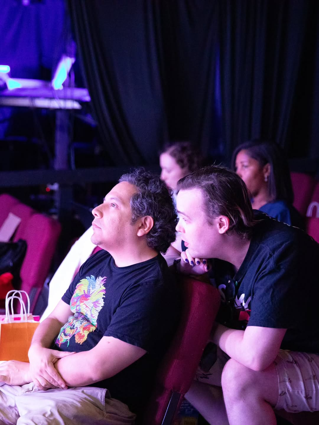 Two men sitting in theater seats watching a performance, with others in the background, in a dimly lit venue.