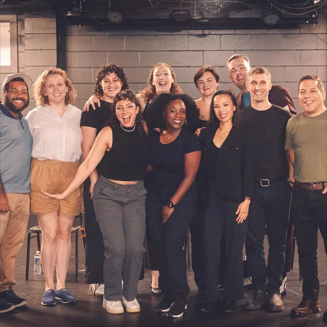 Group of 12 diverse people smiling and posing together on a stage or in a studio.
