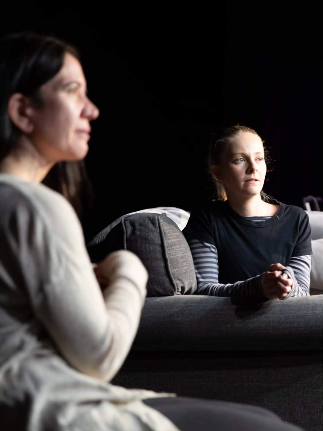 Two women sitting on a couch in a dark room, with one woman in the foreground blurred and the other woman in the background looking serious with hands clasped.