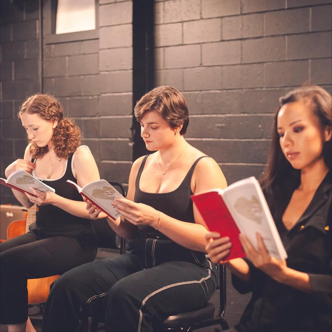 Three women sitting and reading scripts or books in a dimly lit room with dark brick walls.