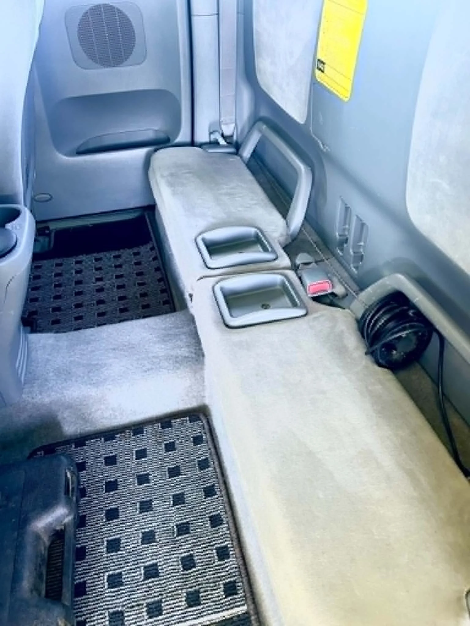 An airplane seating area showing a gray seat with a gray headrest cover, a black and gray patterned floor mat, and part of the airplane interior including a gray wall, a yellow safety instruction sign, and a black air vent.