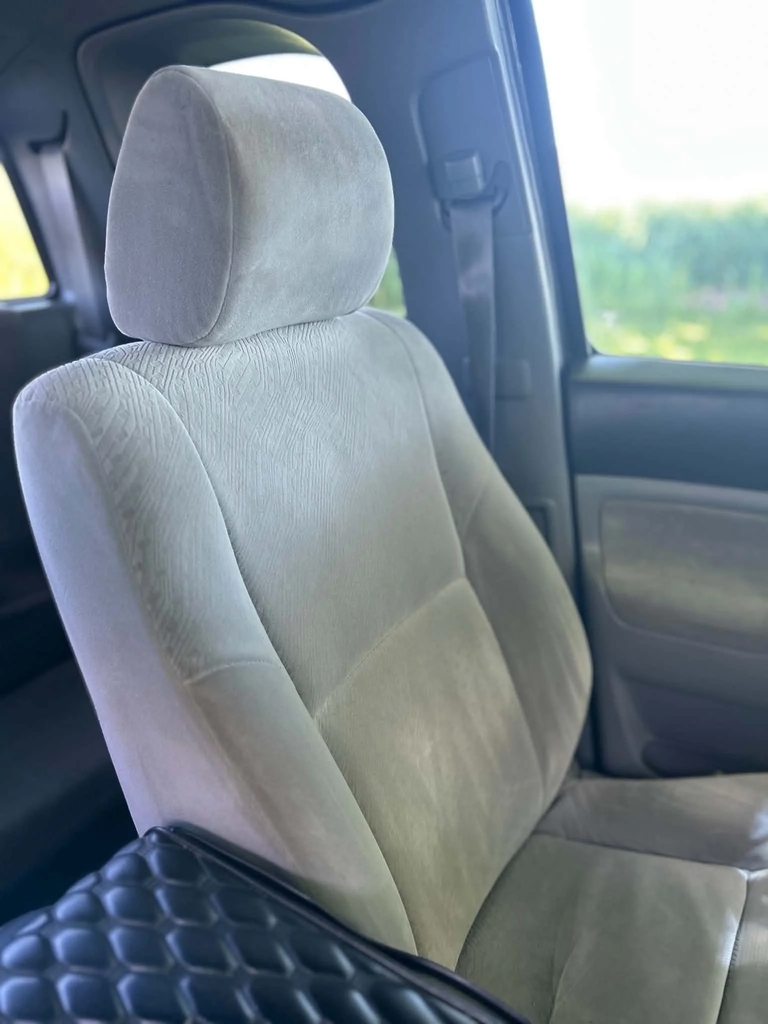 The interior of a vehicle showing the back of a light-colored fabric car seat with an adjustable headrest, and a window revealing a green outdoor landscape.