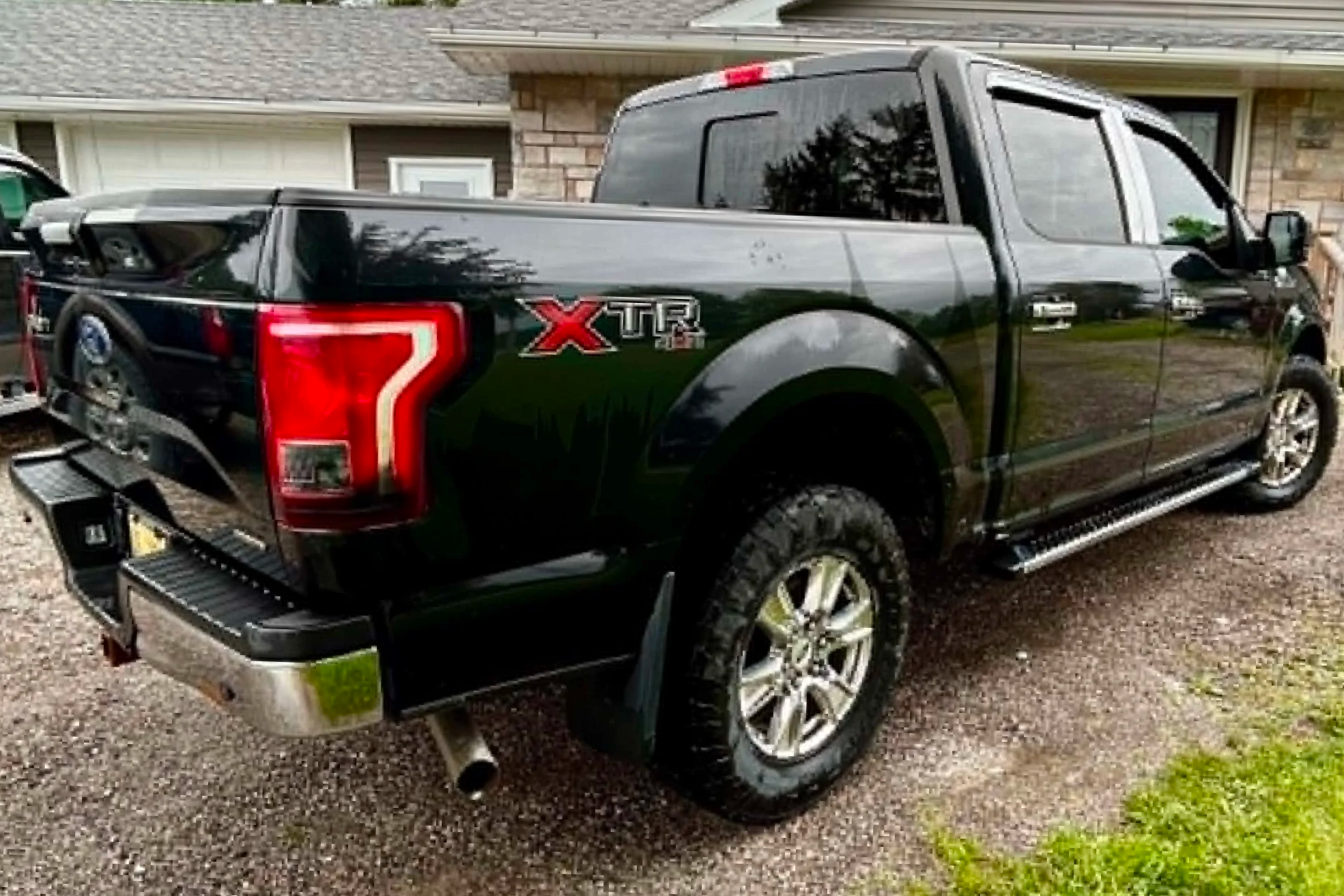A black Ford pickup truck parked in a gravel driveway in front of a house with a brick and siding exterior. The truck has a crew cab with tinted windows and a short bed. There is a damaged rear side panel of the truck.