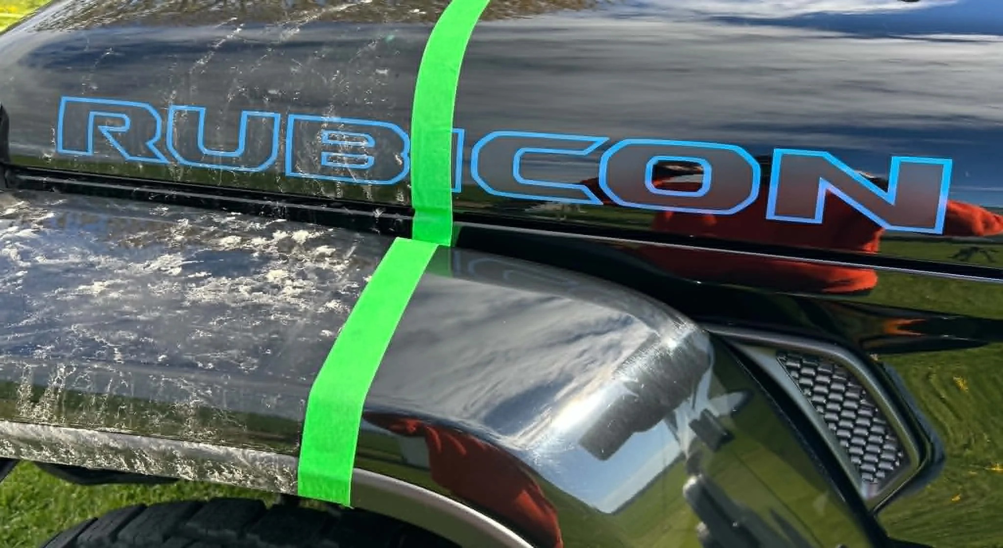 Close-up of a shiny black car with a green stripe, displaying a decal that reads 'RUBICON' in blue outlined letters on the side panel and a red vehicle reflected in the surface.