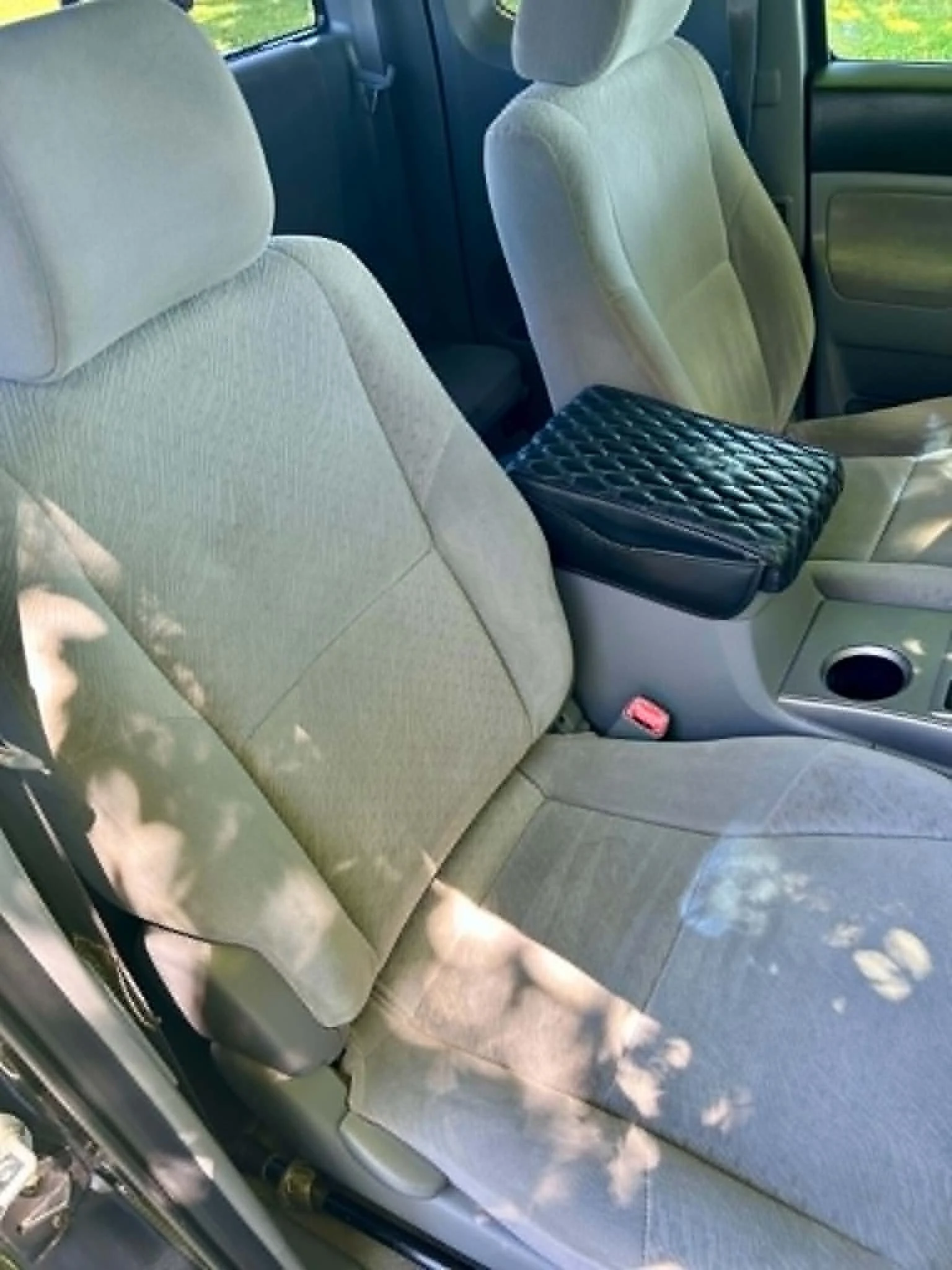The interior of a vehicle showing beige cloth front seats, with a quilted black purse placed on the center console, and shadows of trees on the seats and floor.
