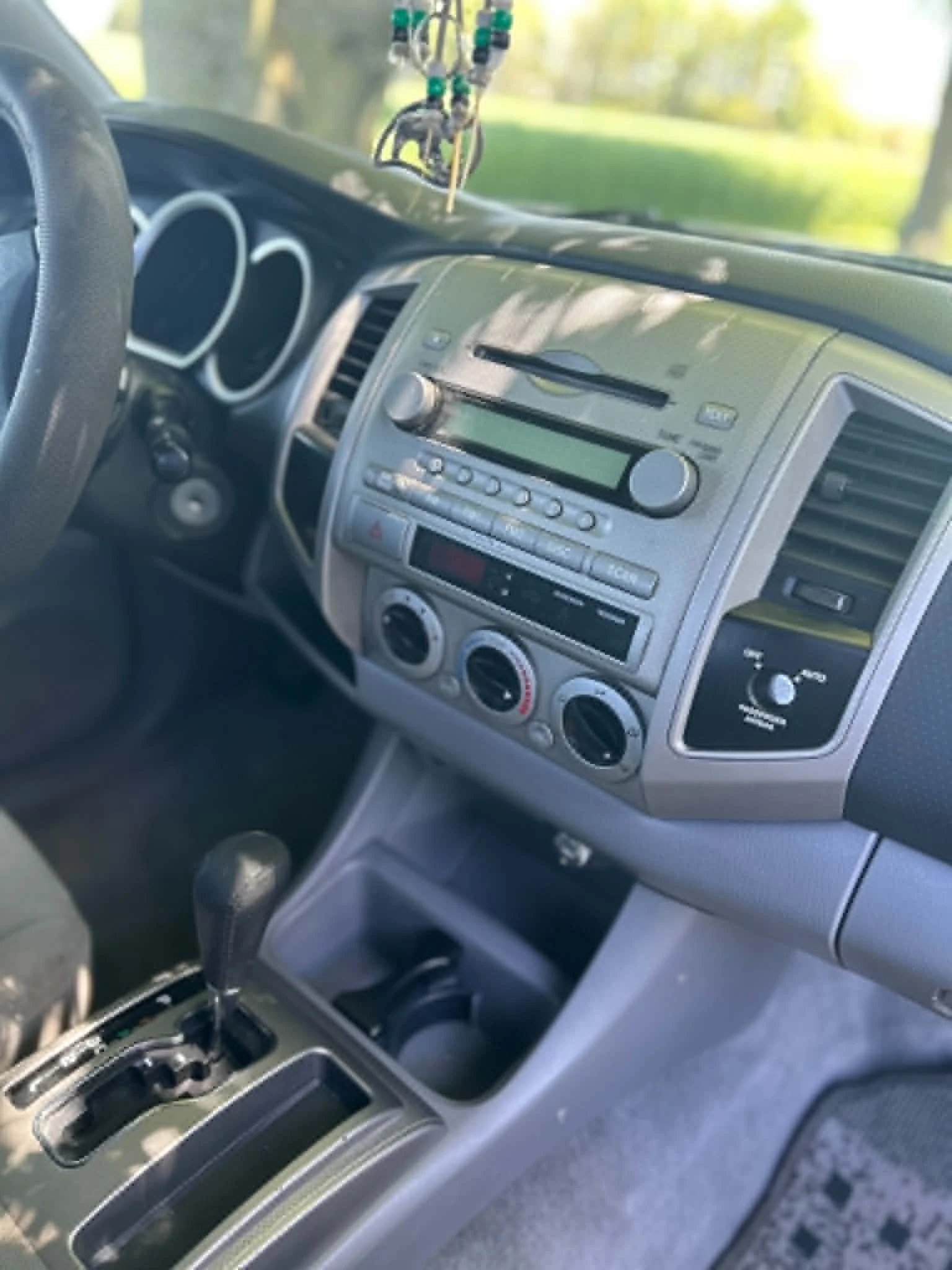 The image shows the interior of a car, focusing on the center console with a gear shifter, climate controls, and an audio system. The dashboard and part of the steering wheel are visible, with a hanging rosary above. The background outside the car indicates a sunny day with trees and grass.