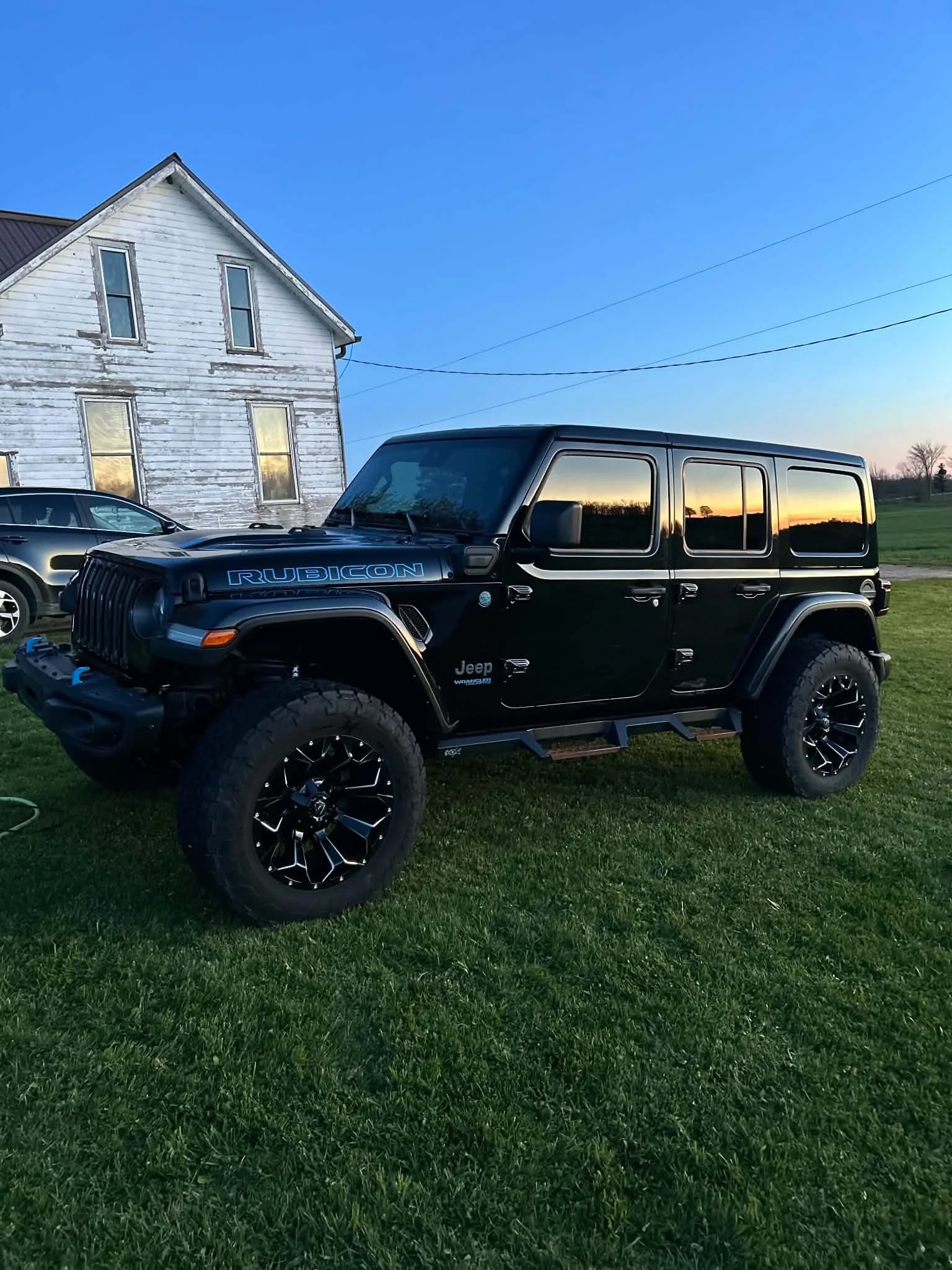Black Jeep Rubicon parked on grass at sunset with a white house in the background and a clear blue sky.