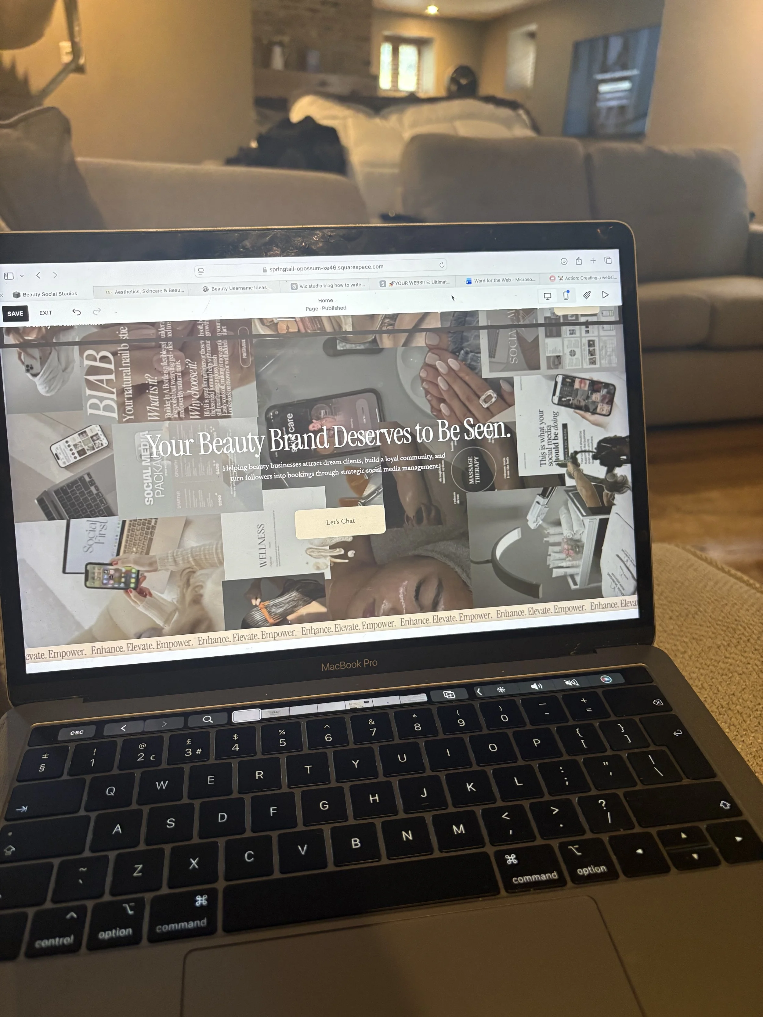 Close-up of a MacBook Pro laptop keyboard with a partially visible screen displaying a website about beauty branding and social media marketing, with a living room visible in the background.