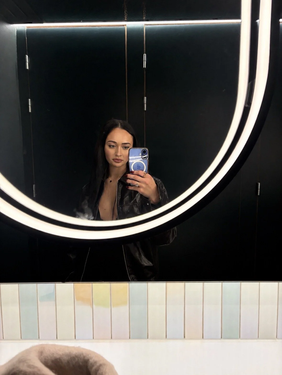 A woman with long dark hair taking a selfie in a mirror with a ring light in a bathroom with black walls and tiled mirror edge.