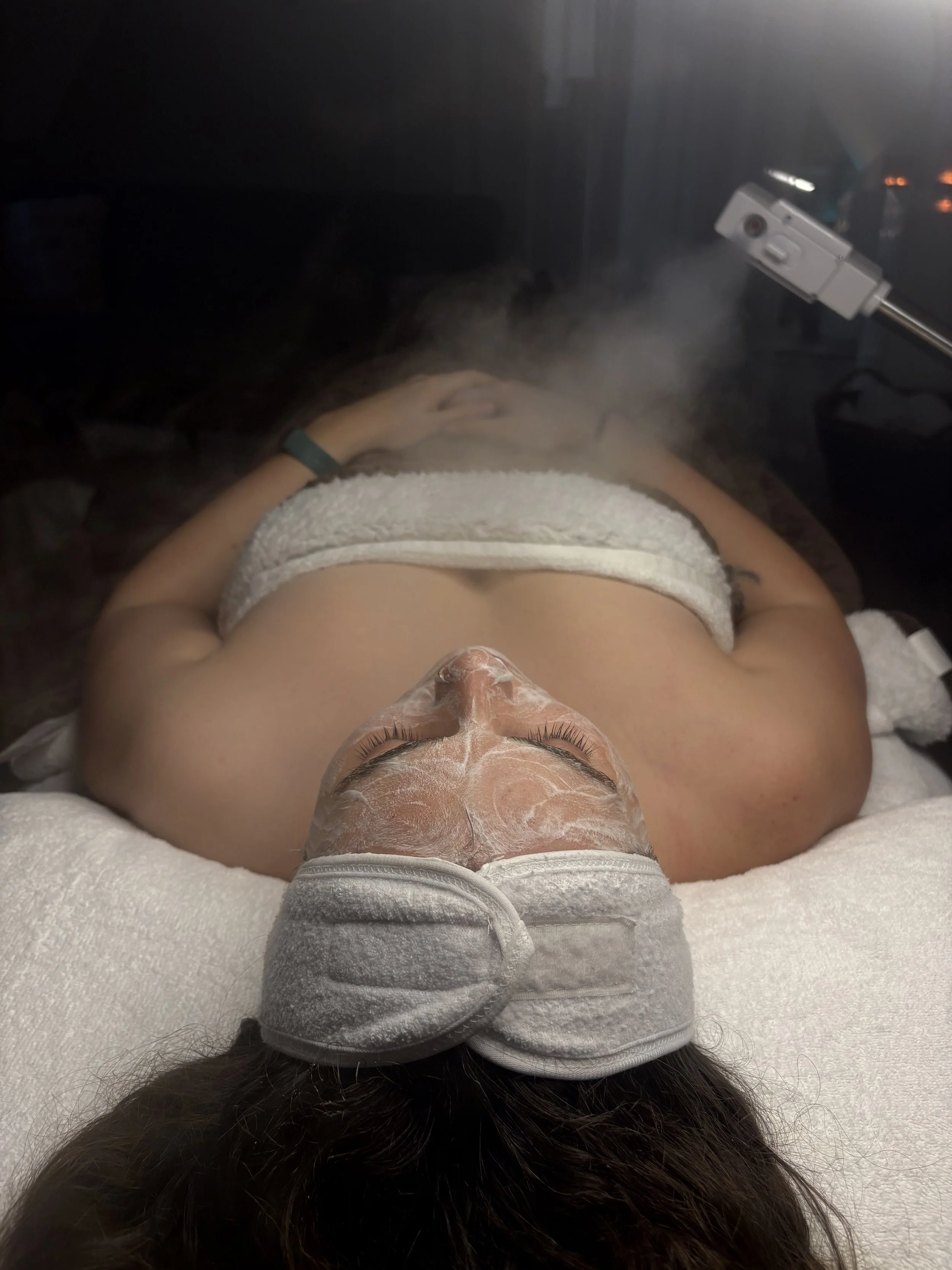 A person lying on a bed with a towel wrapped around their head, receiving a facial treatment. They have a facial mask applied to their face and are wearing protective goggles. Steam emanates from a machine directed at their face.
