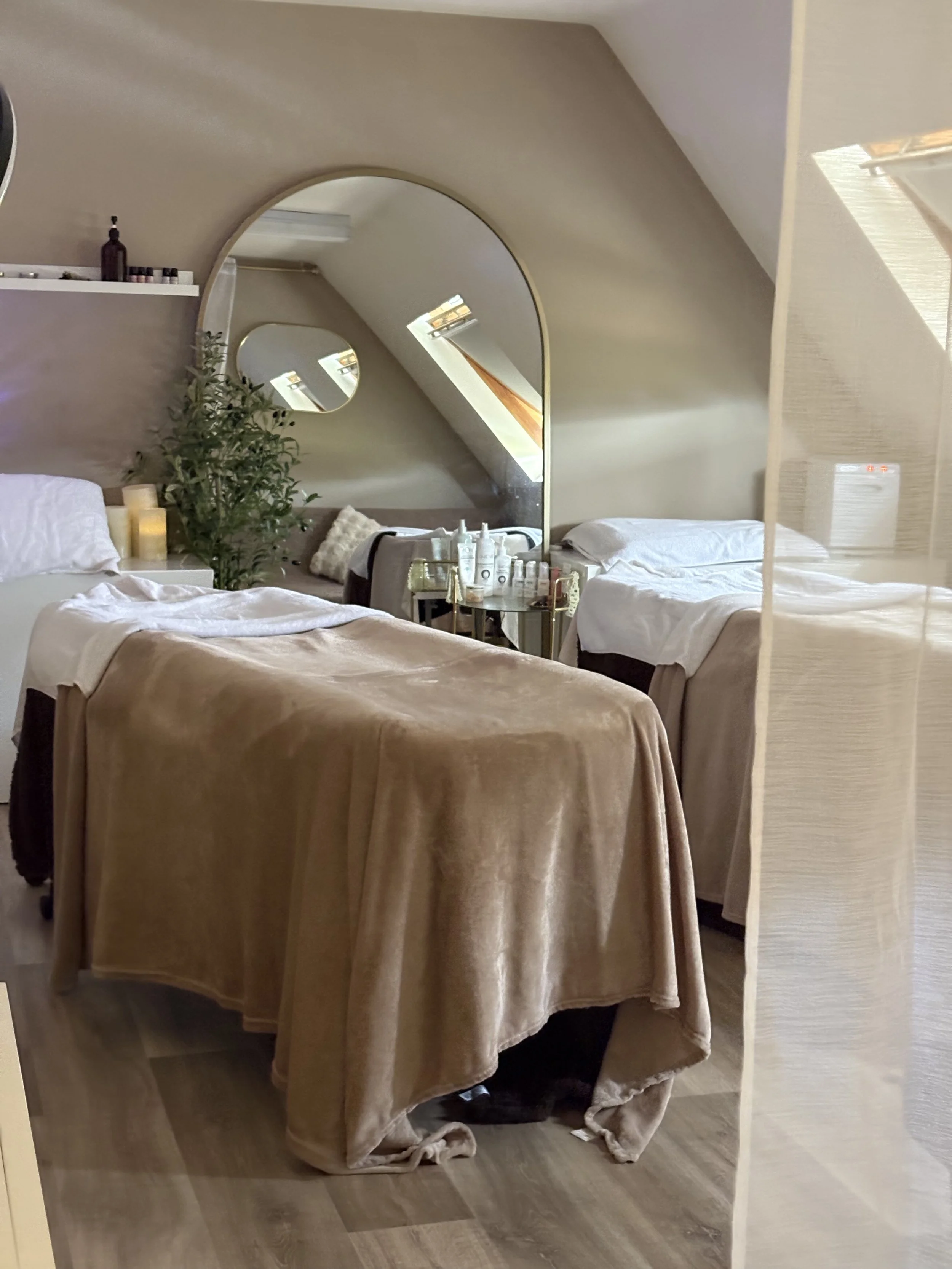 A bedroom with two beds covered in beige bedding, a white shelf with small bottles and candles, a large round mirror on the wall, a smaller mirror on the window, and a side table with skincare products. There is a plant and candles on the side table.