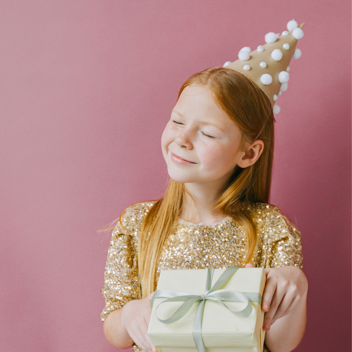 A young girl with red hair smiling with her eyes closed, wearing a gold sequined dress and a cone-shaped party hat decorated with white pom-poms and pearls, holding a gift box wrapped in cream paper with a silver ribbon, against a pink background.