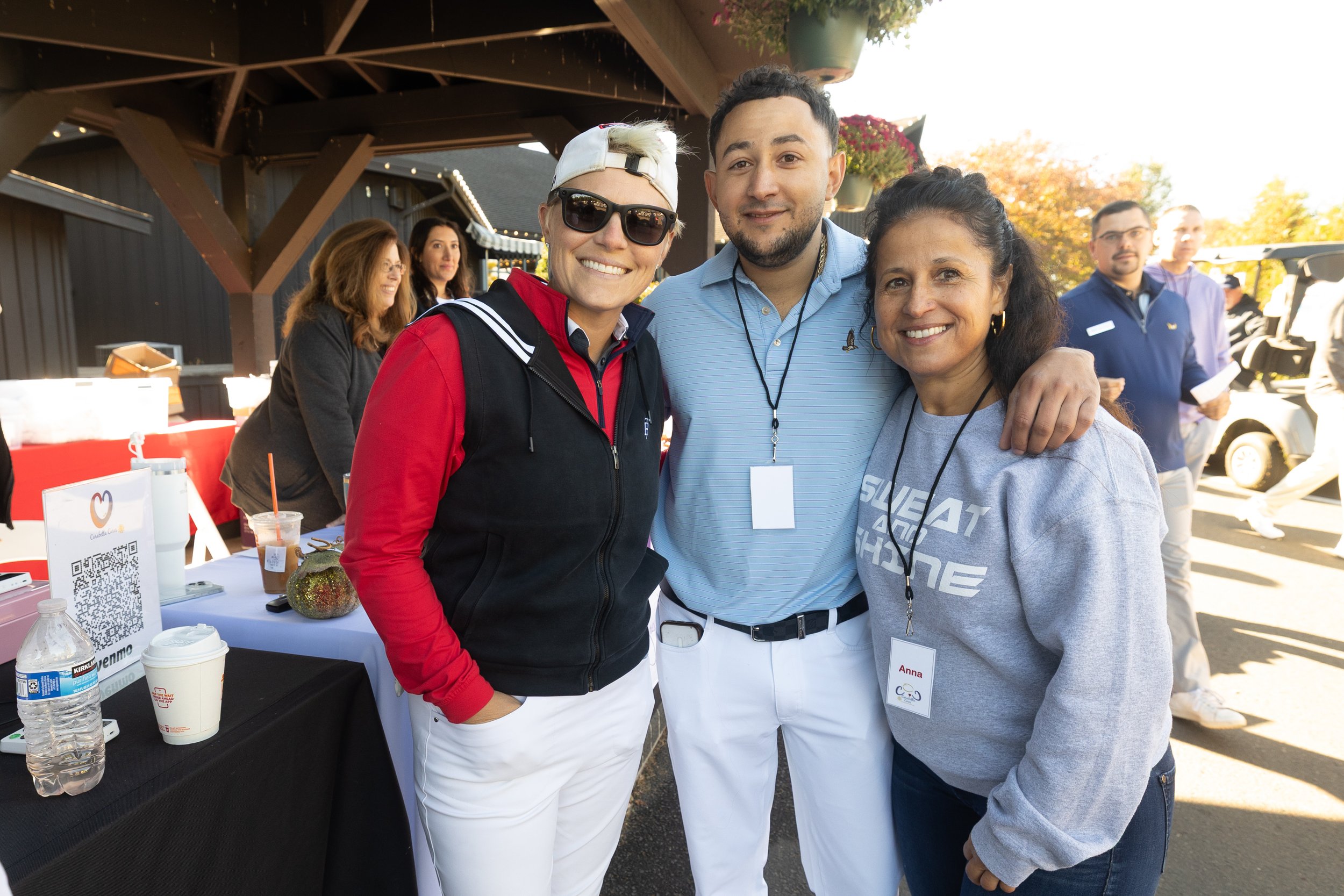 Group of three smiling people posing outdoors at an event, with three other people and event booths in the background.