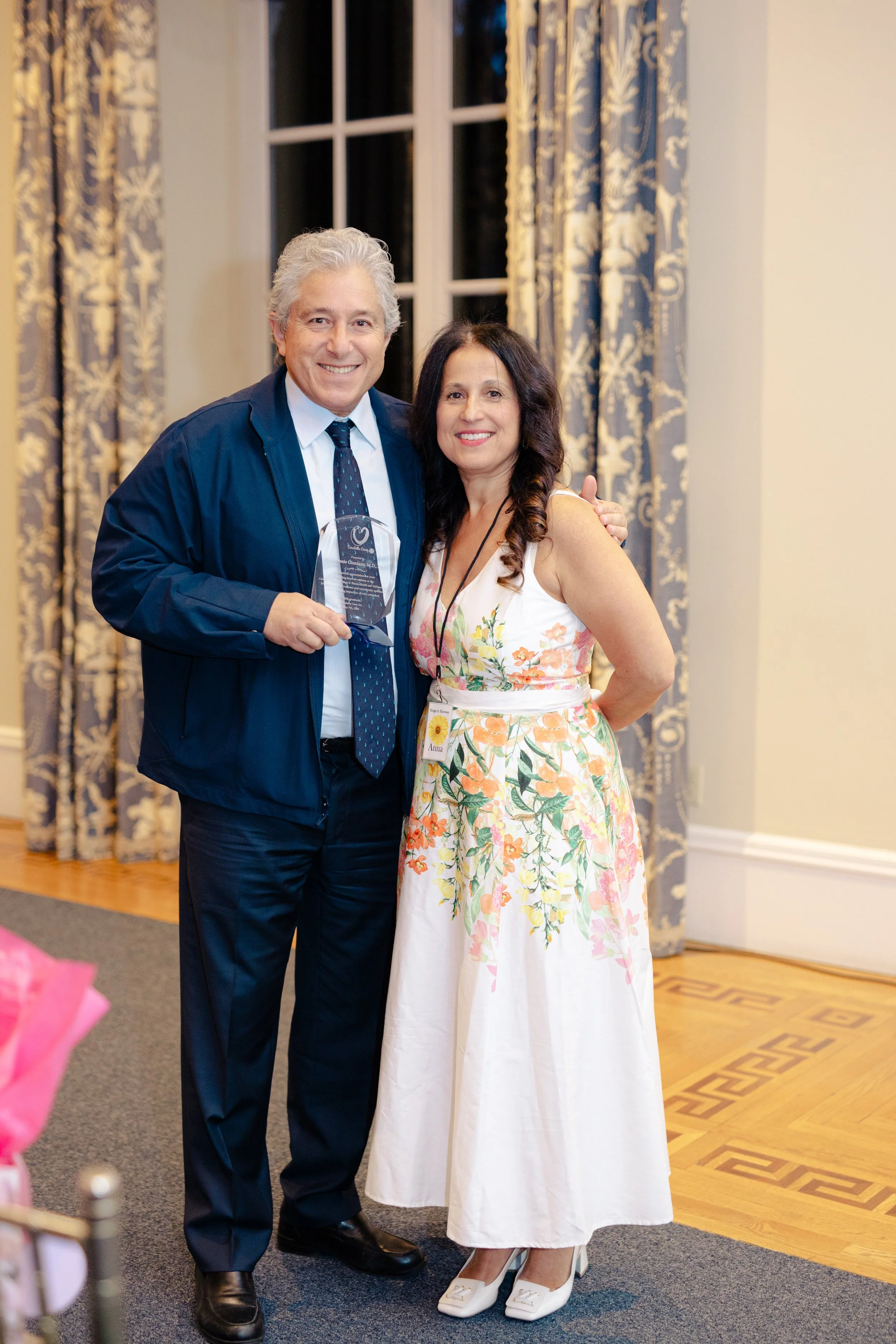 A man in a blue jacket and tie holding a glass award, standing next to a woman in a floral dress. They are smiling and posing indoors, with patterned curtains and a window behind them.