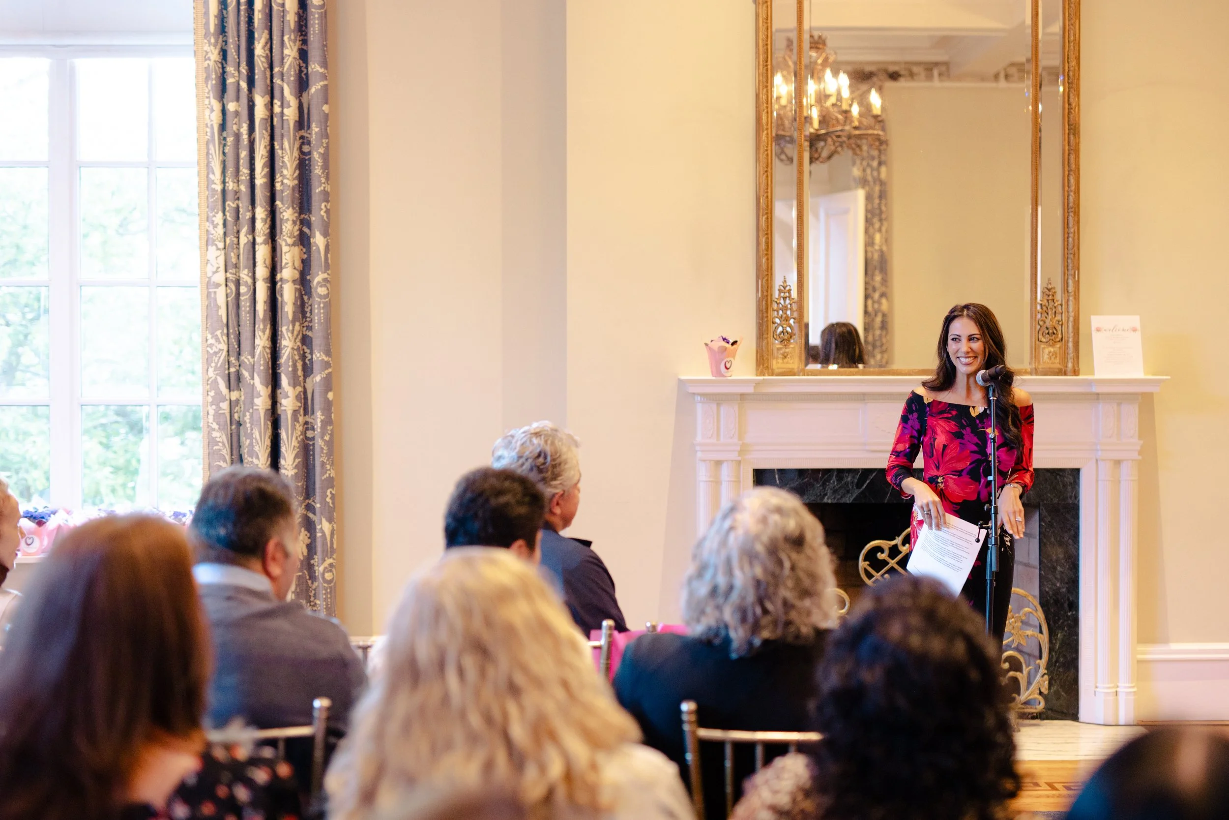A woman with long dark hair, wearing a black and pink floral dress, is speaking or presenting in front of an audience in a formal room with elegant decor. There are several people seated, attentively listening. The room features a large mirror with a gold frame, a white fireplace mantel, a decorative chandelier, and blue patterned curtains by large windows.