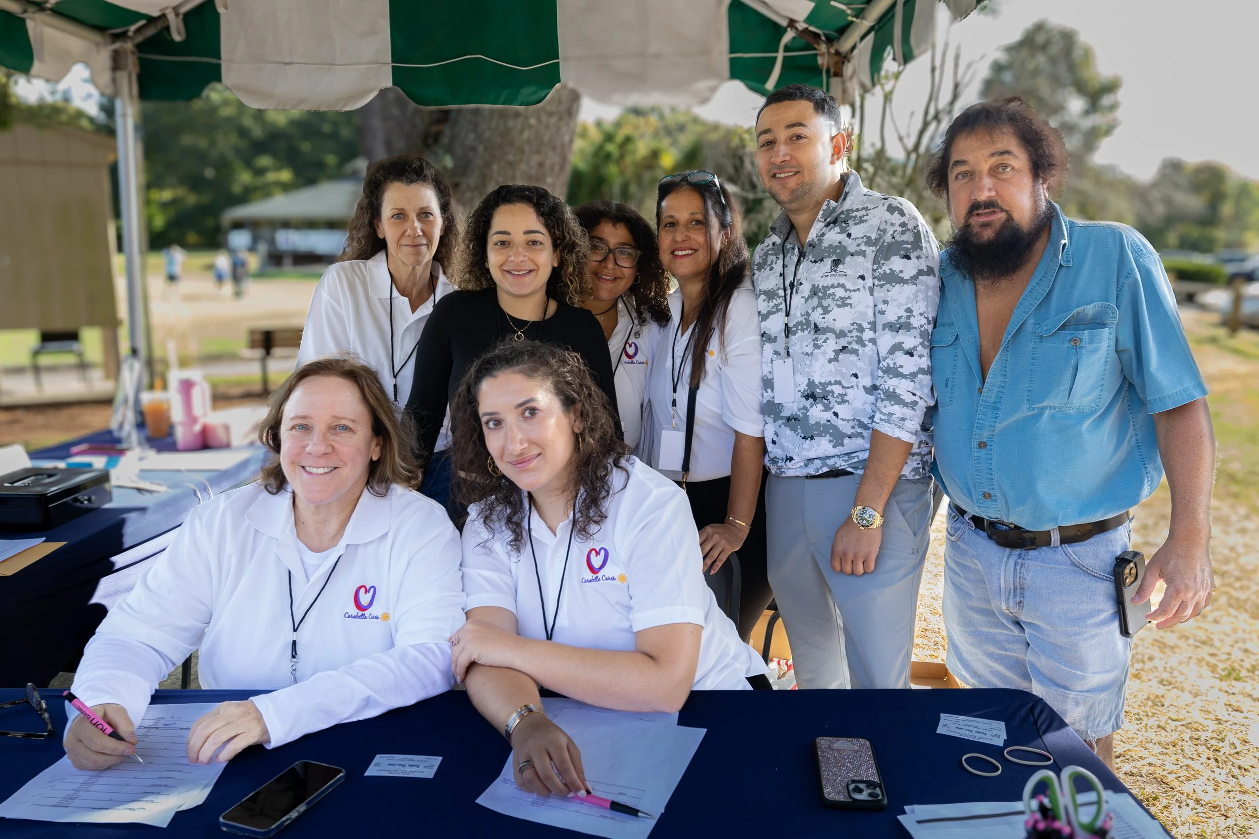 A group of diverse people gathered around a table outdoors, some wearing white shirts with a heart logo, under a canopy, with trees and park scenery in the background.