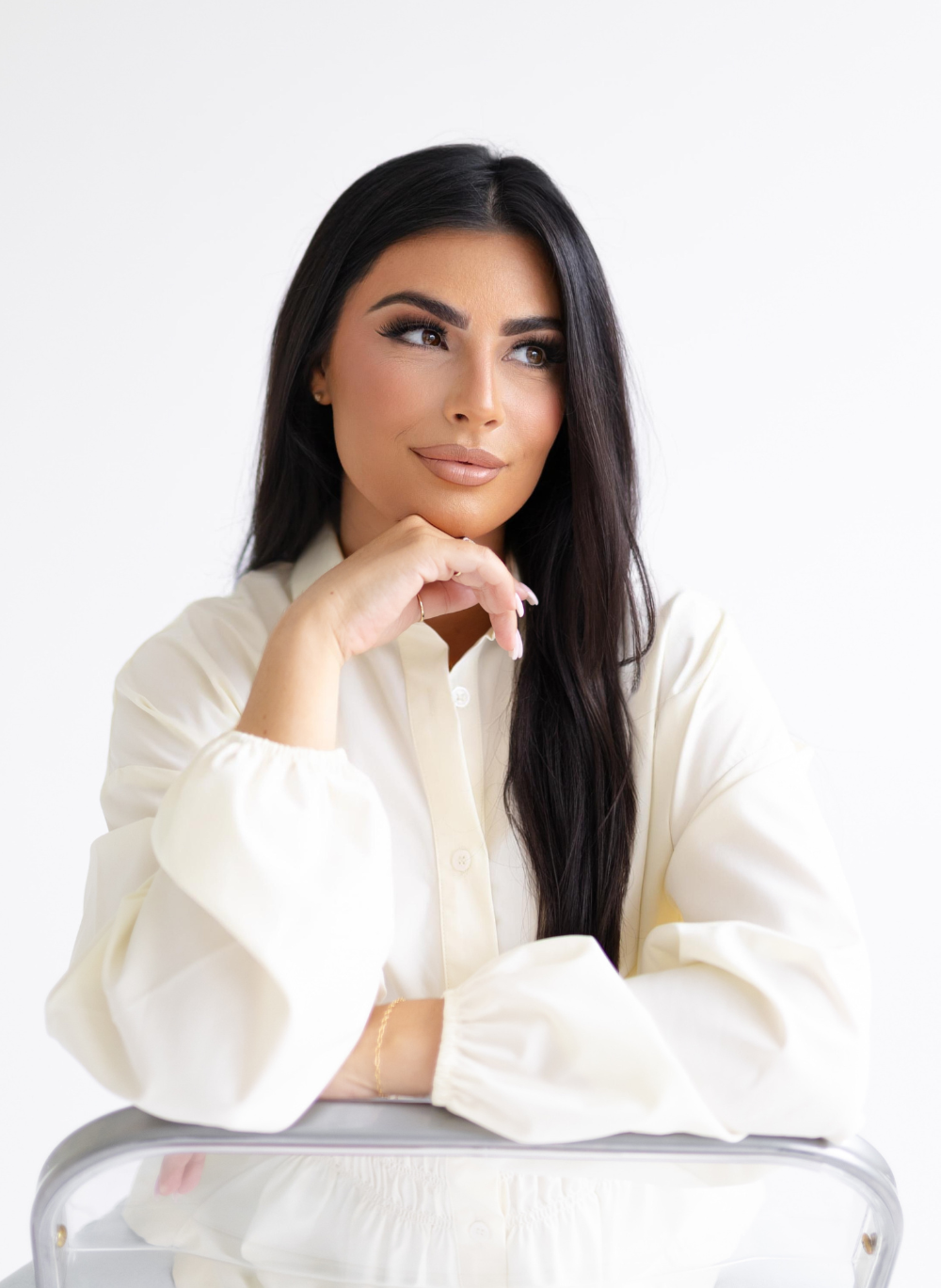 A woman with long dark hair, wearing a cream-colored blouse, is resting her chin on her hand and looking thoughtfully to the side against a plain white background.