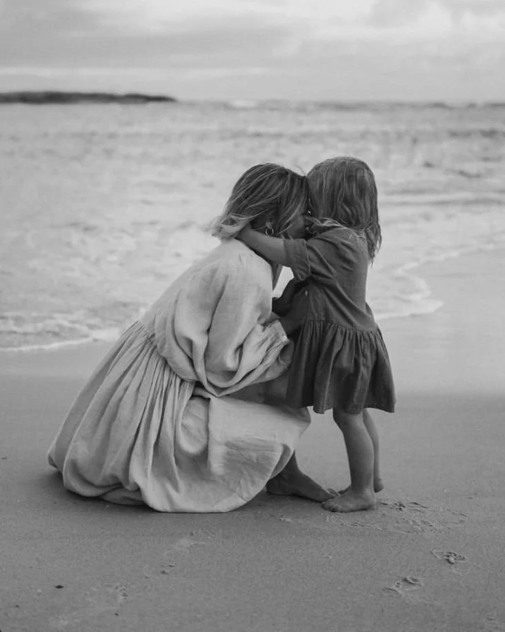 A woman and a young girl share a kiss on the beach, with the woman kneeling and the girl standing on the sand, holding the woman's face.