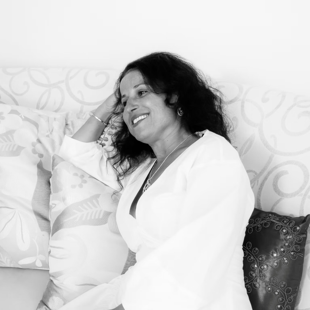 A woman with dark, wavy hair smiling while lying on a bed, resting her head on her hand, surrounded by decorative pillows in a bright room.