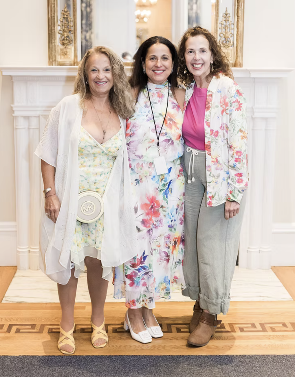 Three women standing together indoors in front of a white fireplace and mirror, smiling at the camera. They are dressed in colorful, floral clothing.