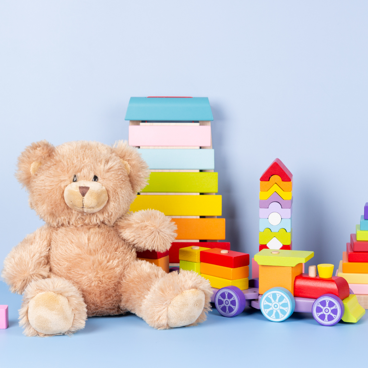 A plush teddy bear sitting on a light blue surface with colorful wooden toys, including blocks, a toy train, and stacking rings, against a light blue background.