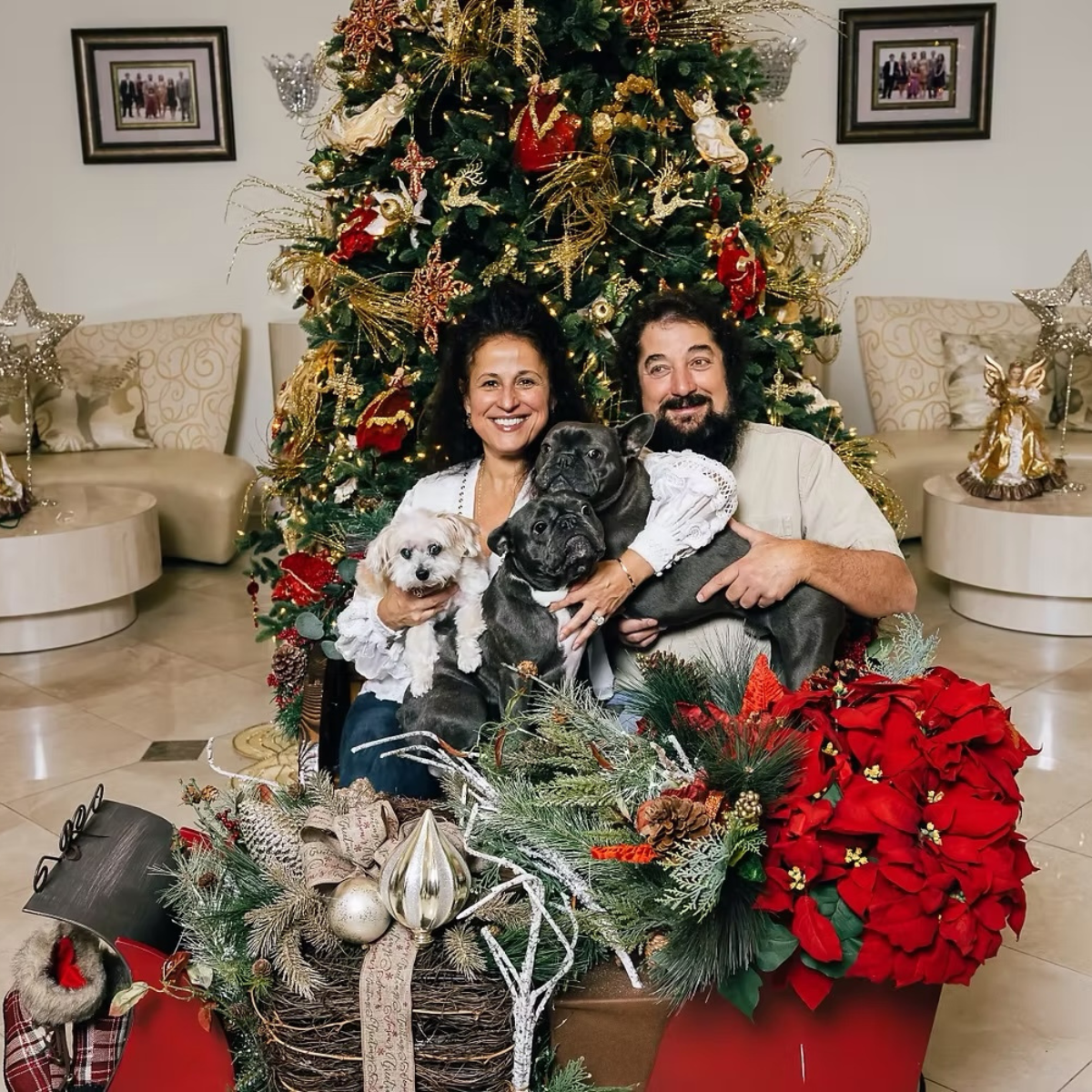 A smiling couple sitting in front of a decorated Christmas tree with three dogs, surrounded by holiday decorations including poinsettias, ornaments, and silver and gold accents.