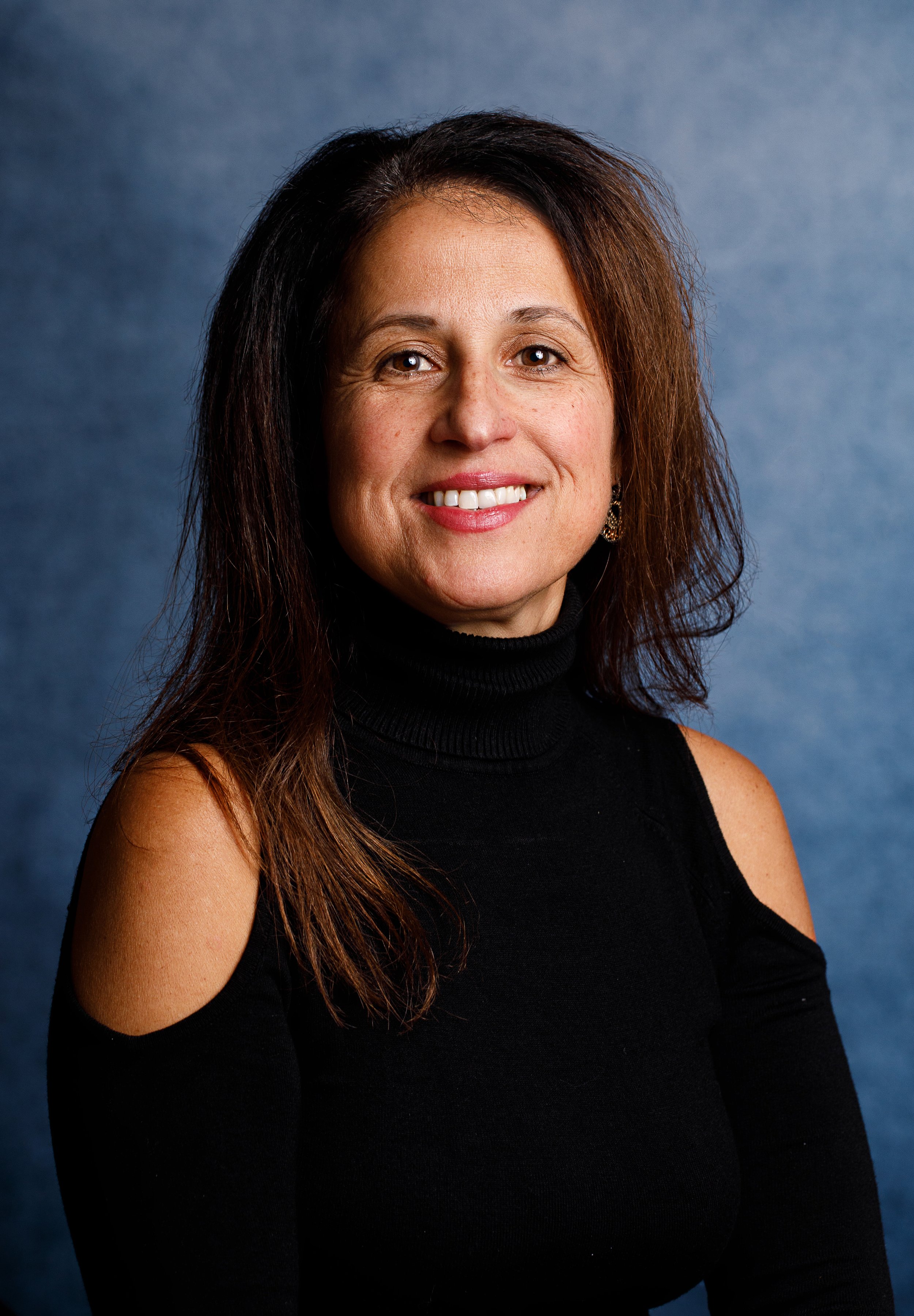 A woman with shoulder-length brown hair, wearing a black top with cut-out shoulders, smiling against a blue background.