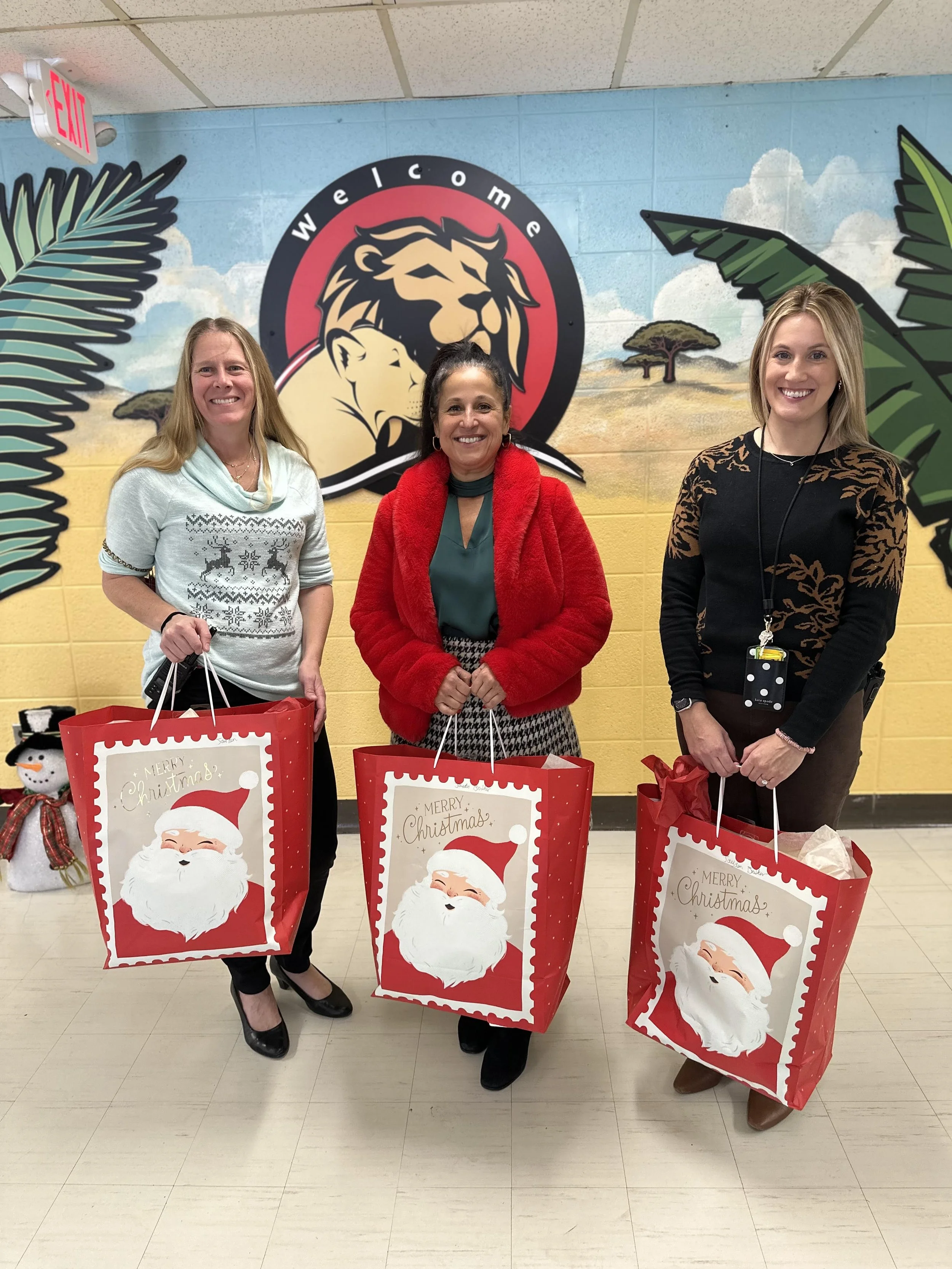 Three women standing indoors at a holiday event, holding large red gift bags with Santa Claus illustrations. The background features a painted mural with a lion and a越zebra in a landscape, along with a large 