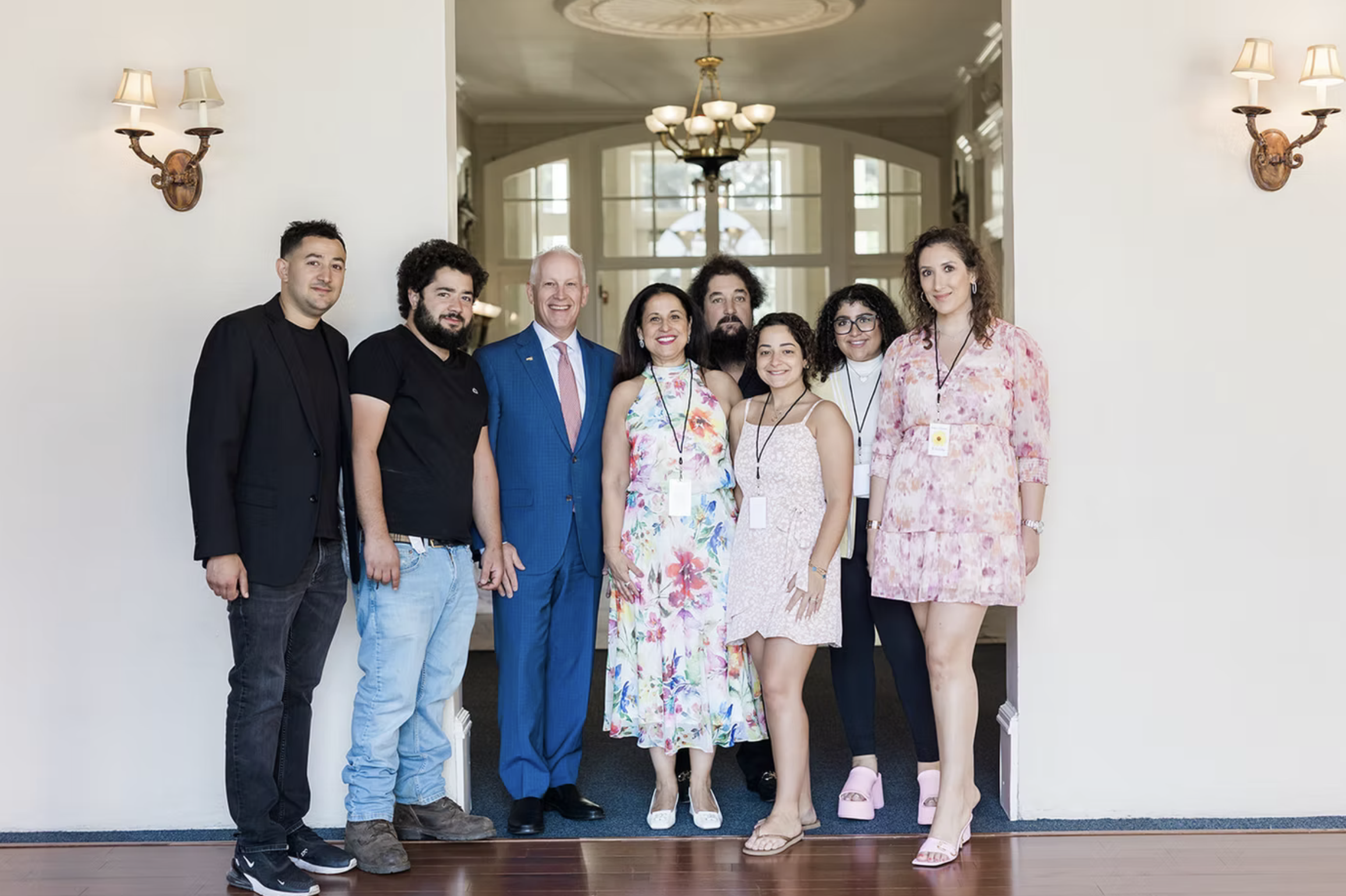 A diverse group of nine people standing together in a hallway. The group includes men and women of different ages and ethnicities. They are smiling and wearing casual and business attire. The background shows large windows, chandeliers, and wall-mounted lamps with fabric shades.