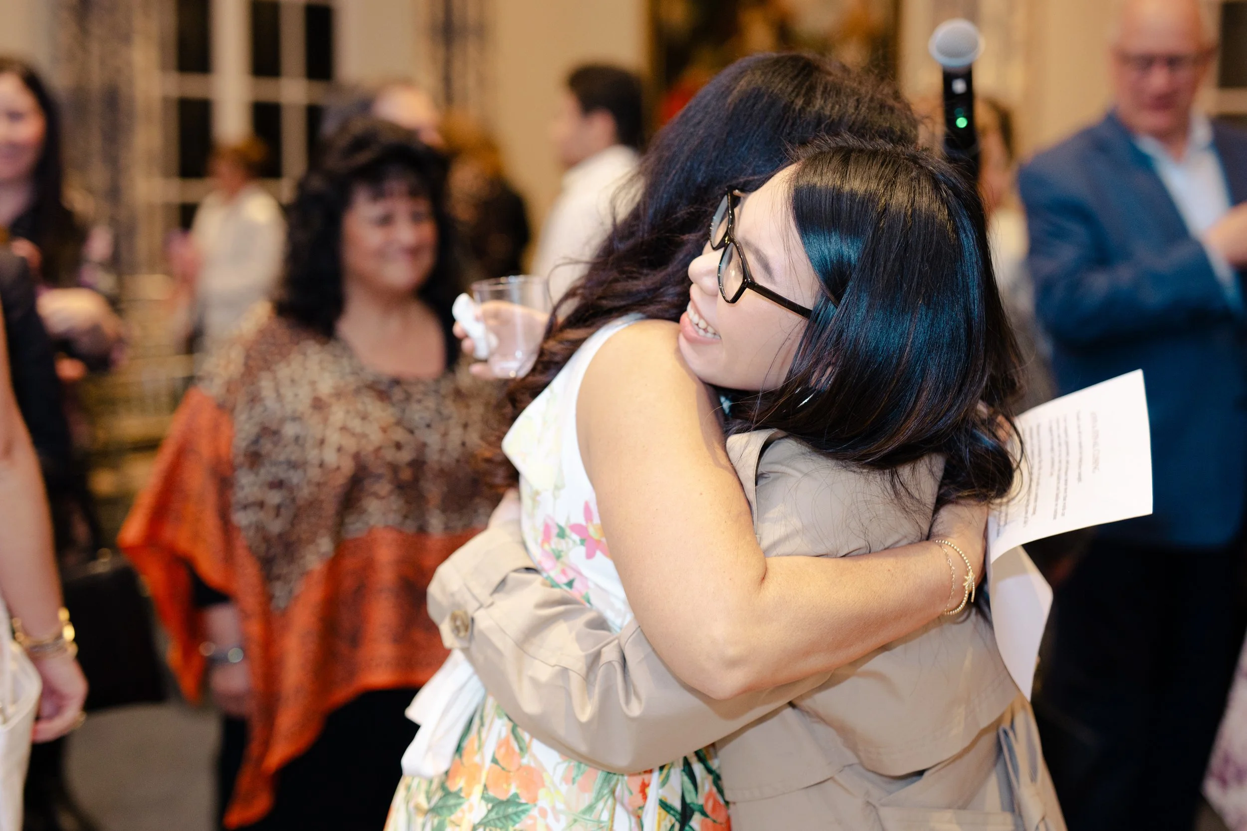 Two women hugging at a social event, surrounded by other people in the background.