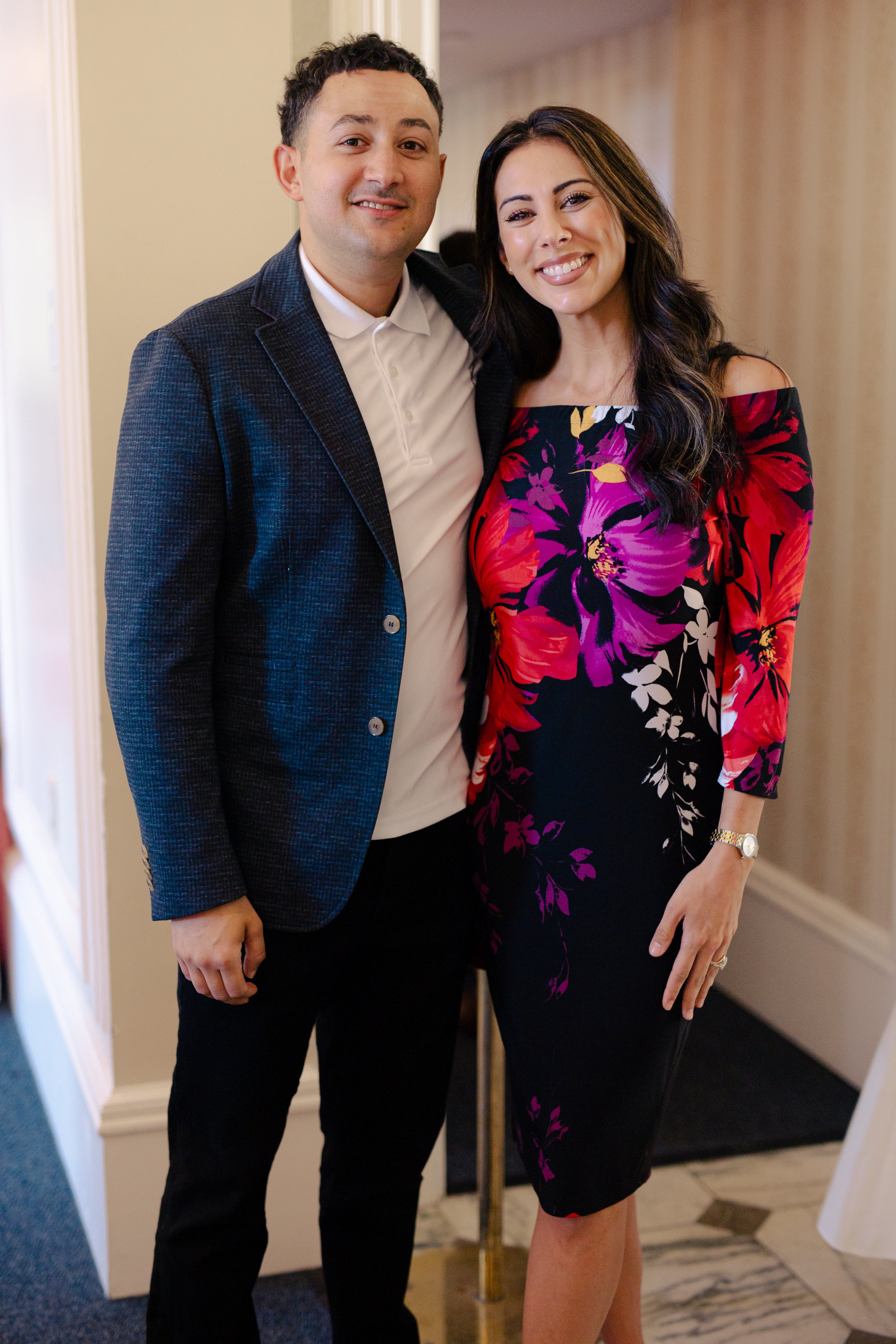 A man and woman stand together indoors, smiling at the camera. The man wears a dark blazer over a white shirt, and the woman wears a colorful off-the-shoulder floral dress, with long dark hair.