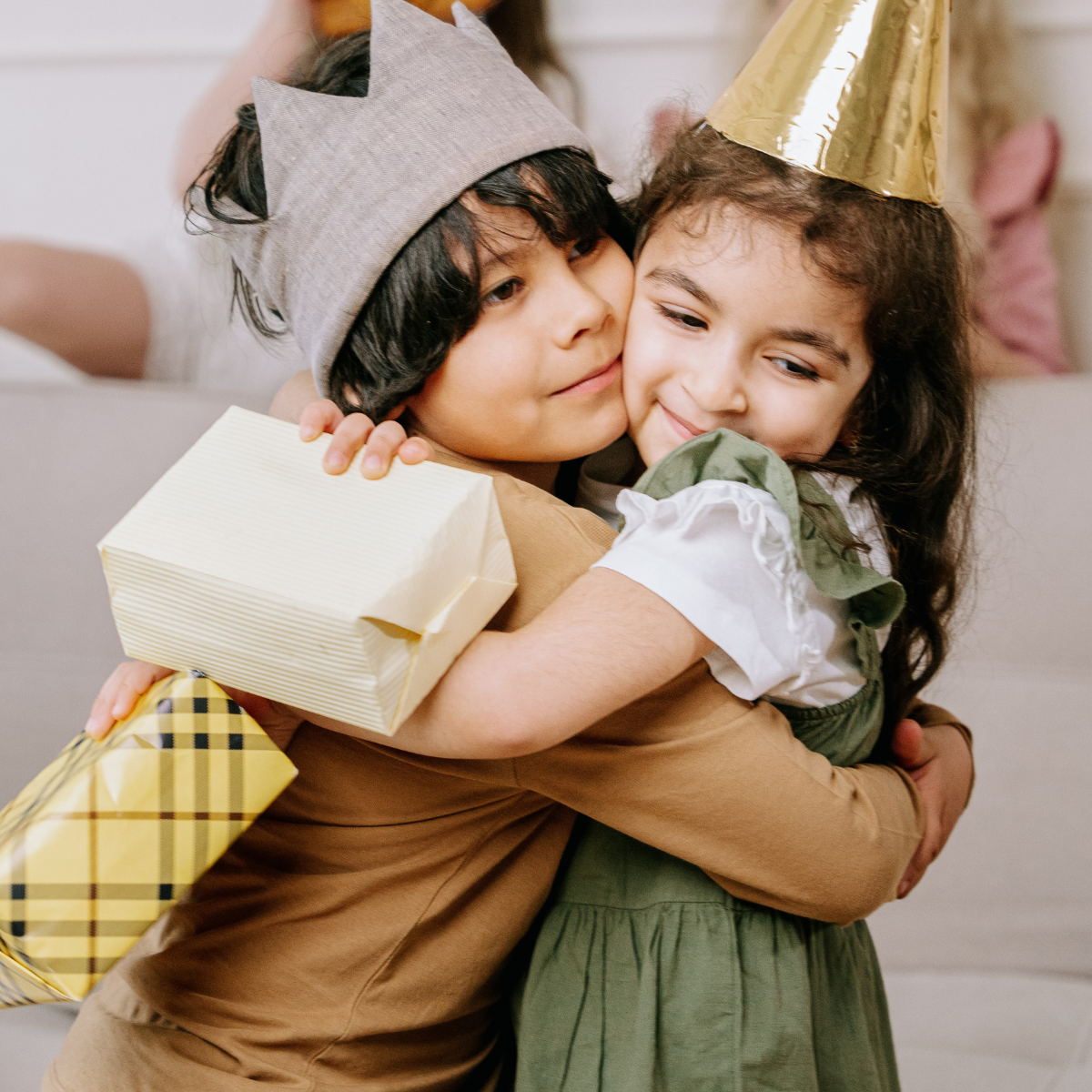 Two kids wearing party hats hugging each other, celebrating a birthday with gifts.