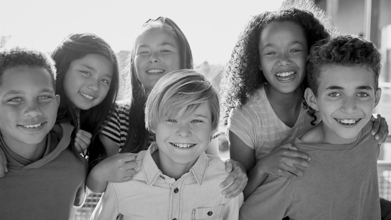 Group of seven smiling children posing together outdoors in bright sunlight.