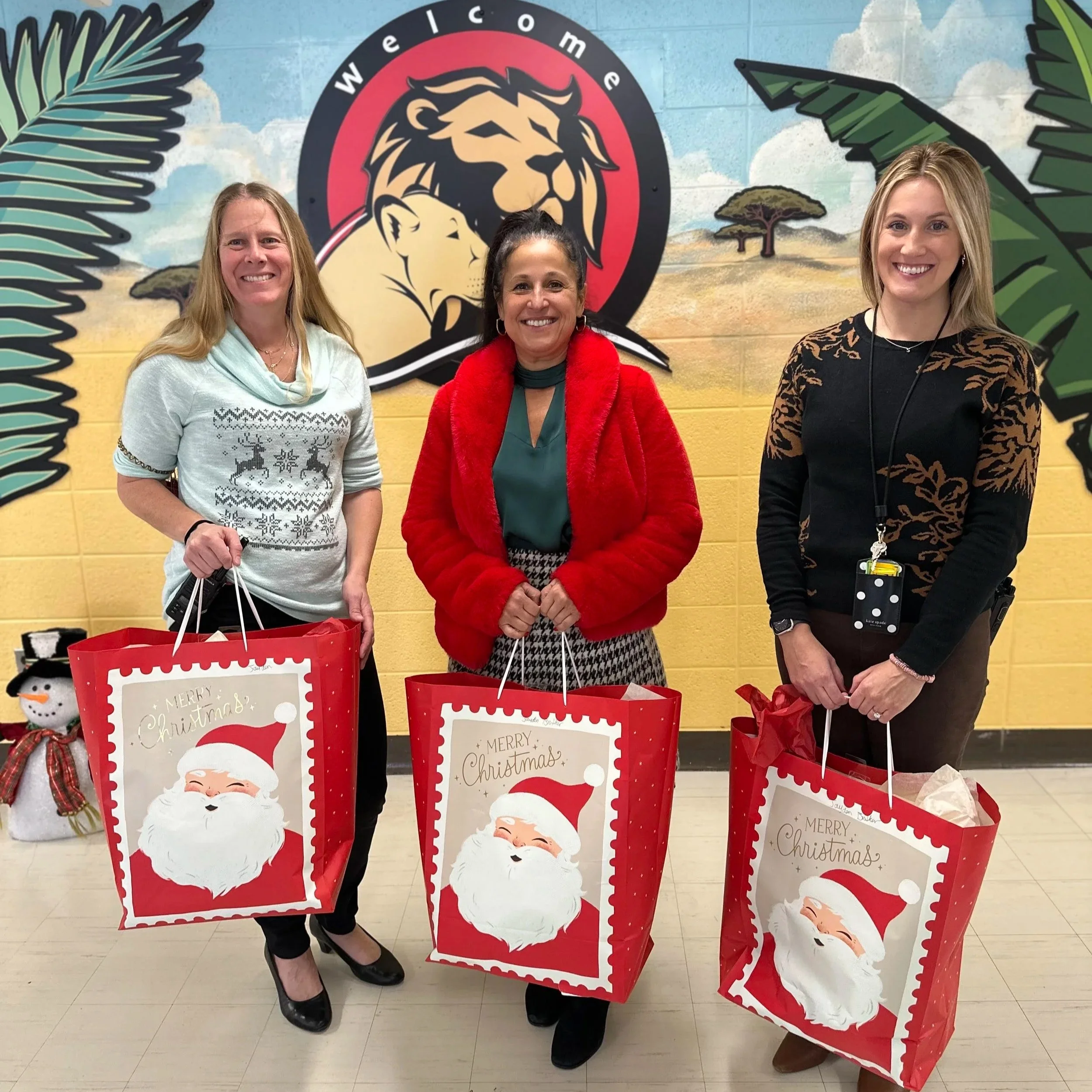 Three women standing indoors in front of a colorful mural with a lion's head, palm leaves, trees, and clouds, holding red gift bags with a Christmas Santa face design and the message 'Merry Christmas'.