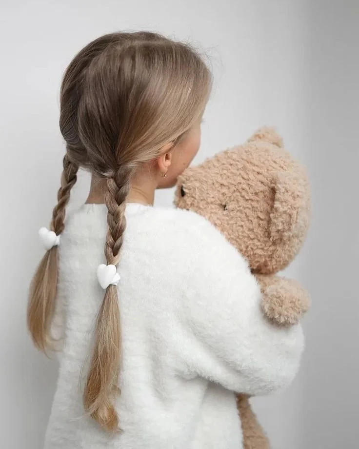 A girl with braided hair holding a teddy bear. She is facing away from the camera and is wearing a white sweater.